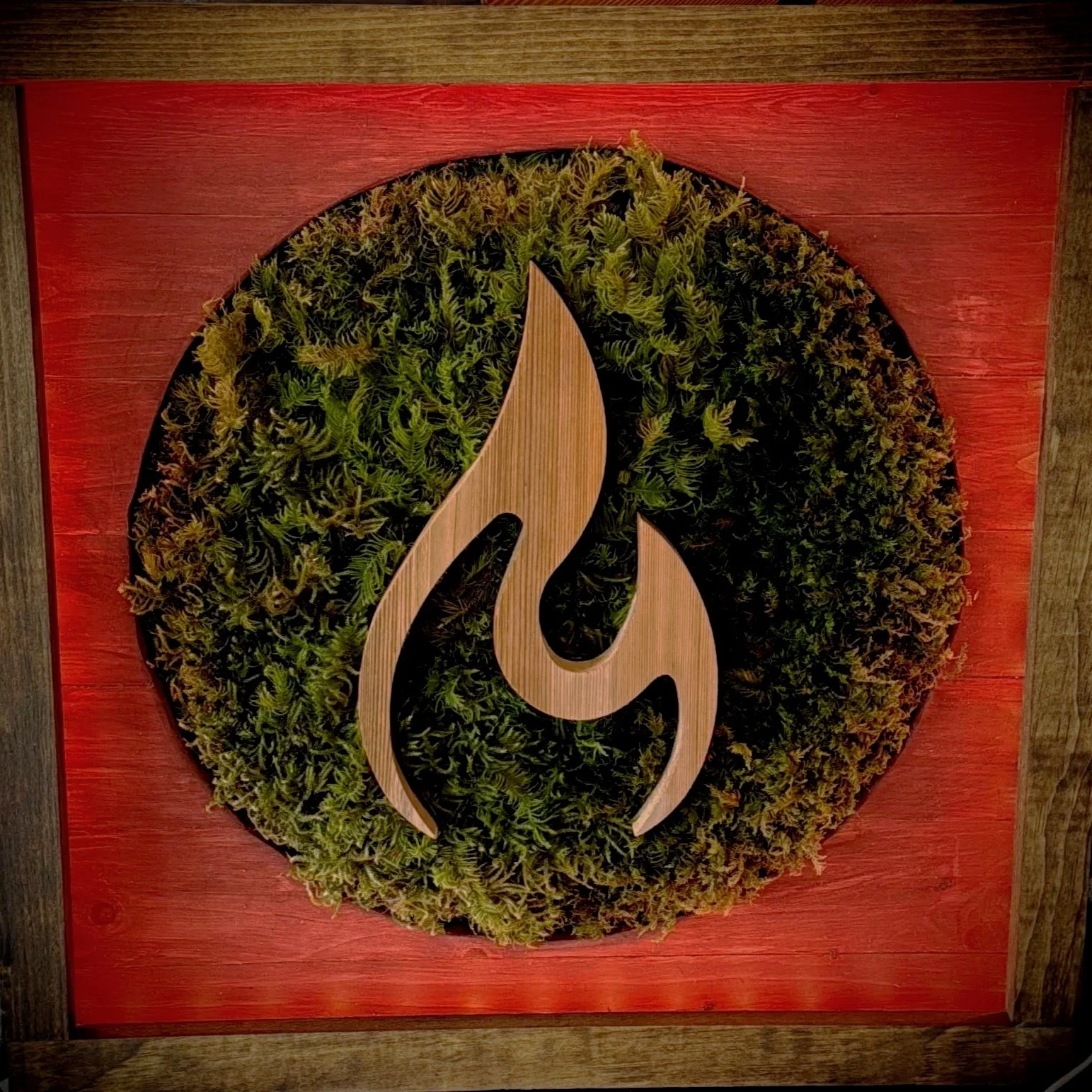 Decorative wall art with a fire-shaped design made of wood, set against a green moss background inside a red wooden frame.