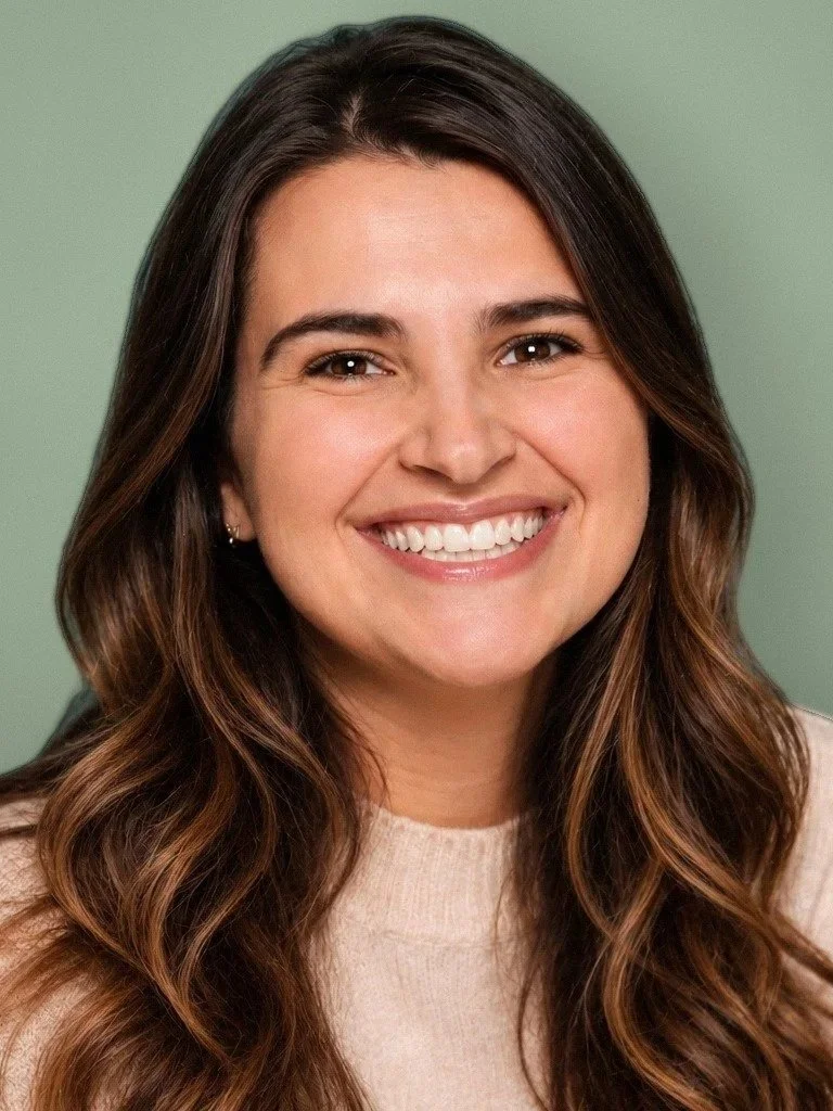 Headshot of a woman with long, wavy brown hair and a bright smile, wearing a beige top, against a muted green background.