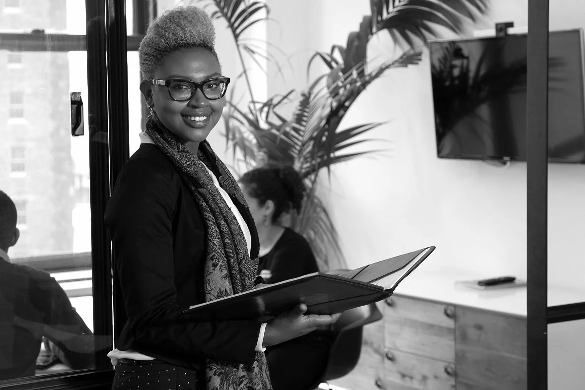 A Black business woman smiling with glasses and a scarf holding a folder in an office.