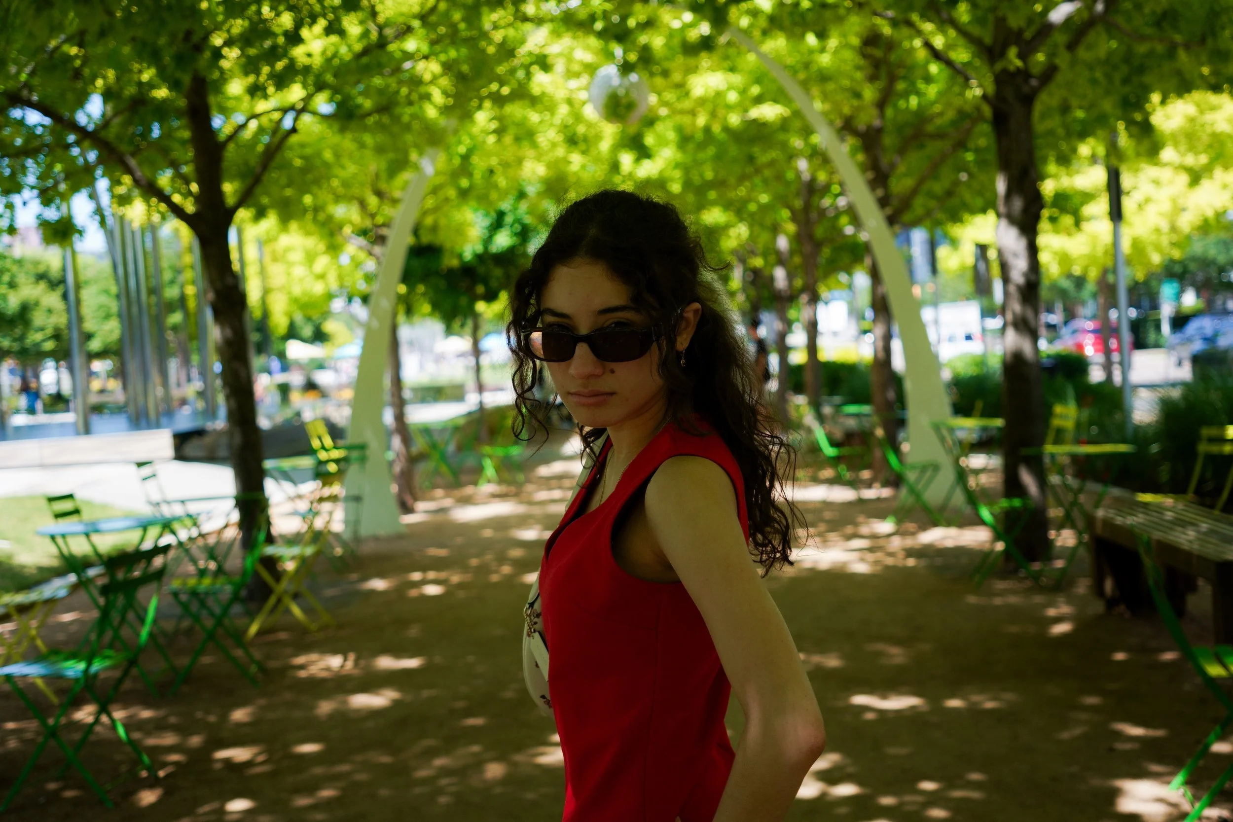 A woman with dark curly hair, wearing sunglasses and a red sleeveless top, standing in a shaded park with green chairs and trees.
