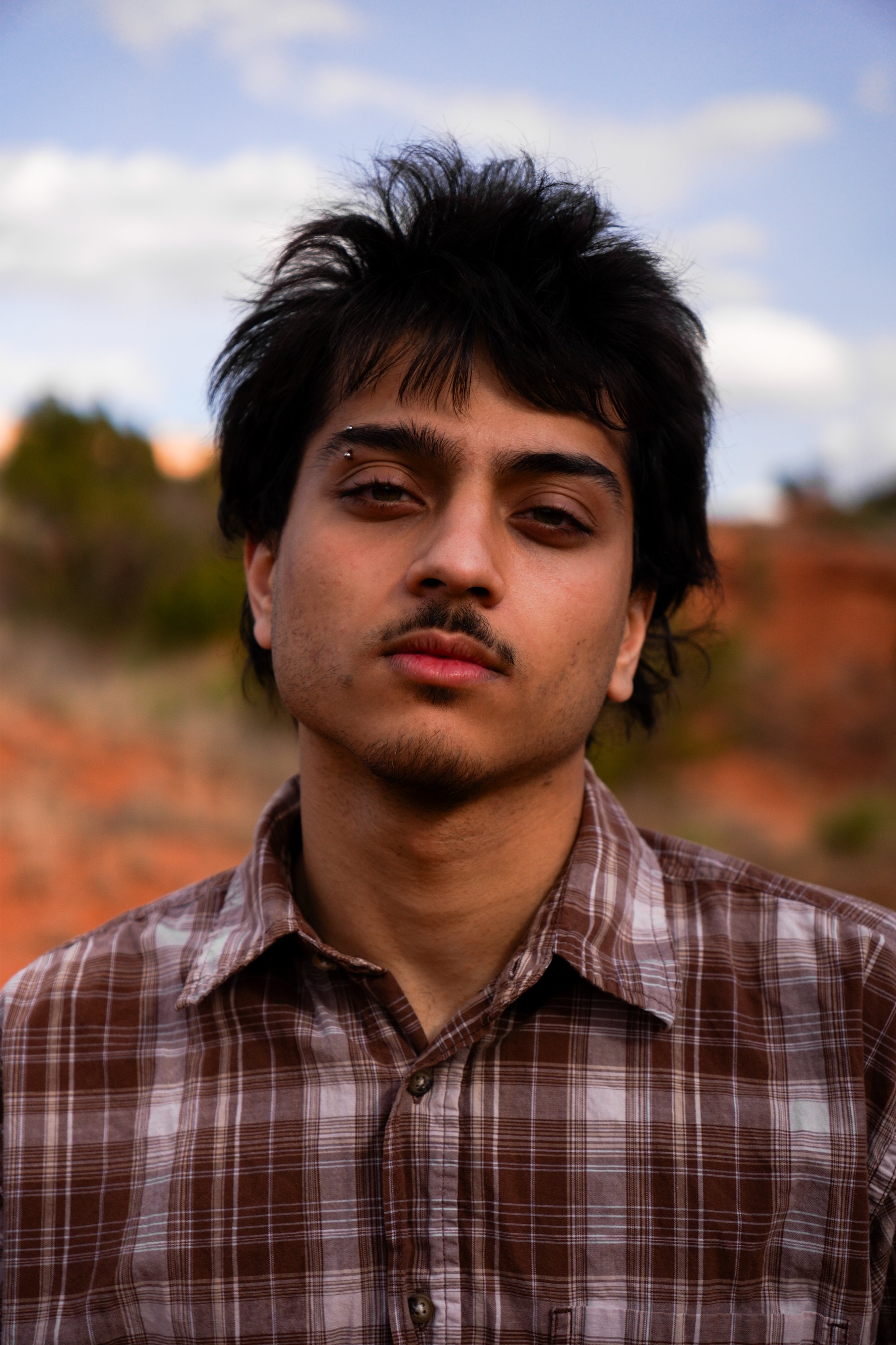 A young man with medium-length black hair, a facial piercing above his right eyebrow, and wearing a plaid shirt, stands outdoors with a blurred natural landscape and partly cloudy sky in the background.