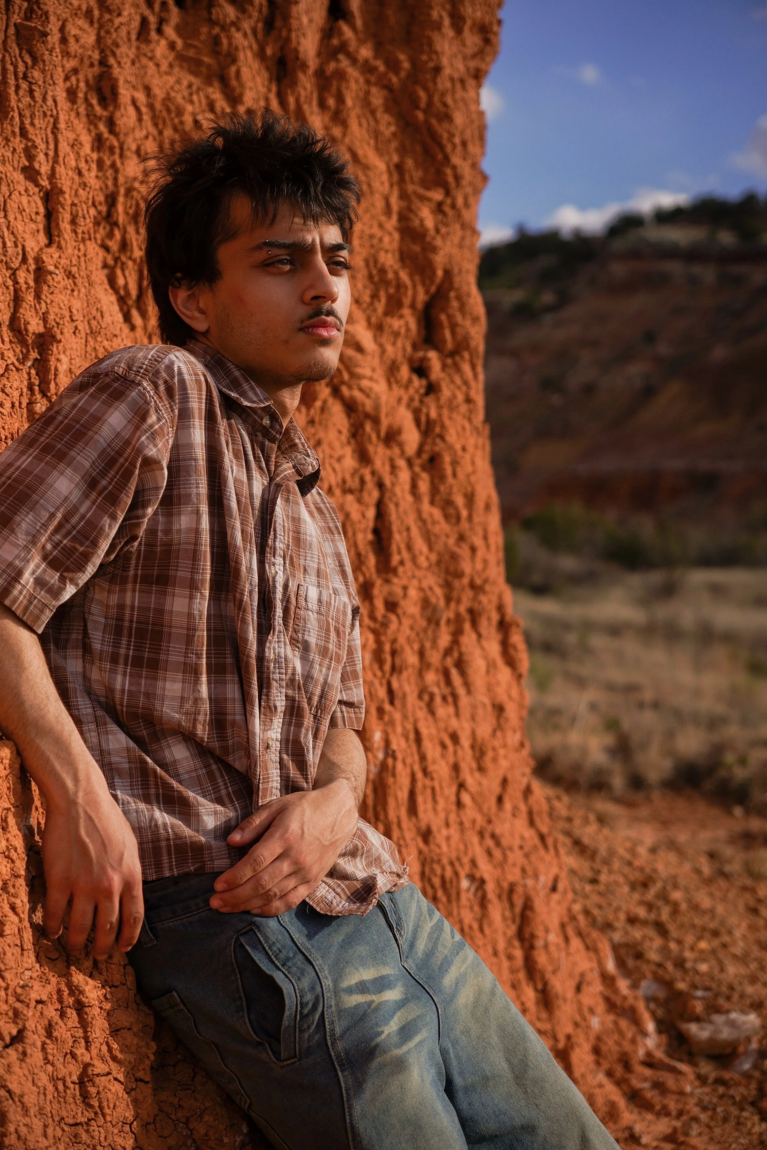 A young man wearing a plaid shirt and jeans leaning against an orange rock formation in an arid landscape under a blue sky with clouds.