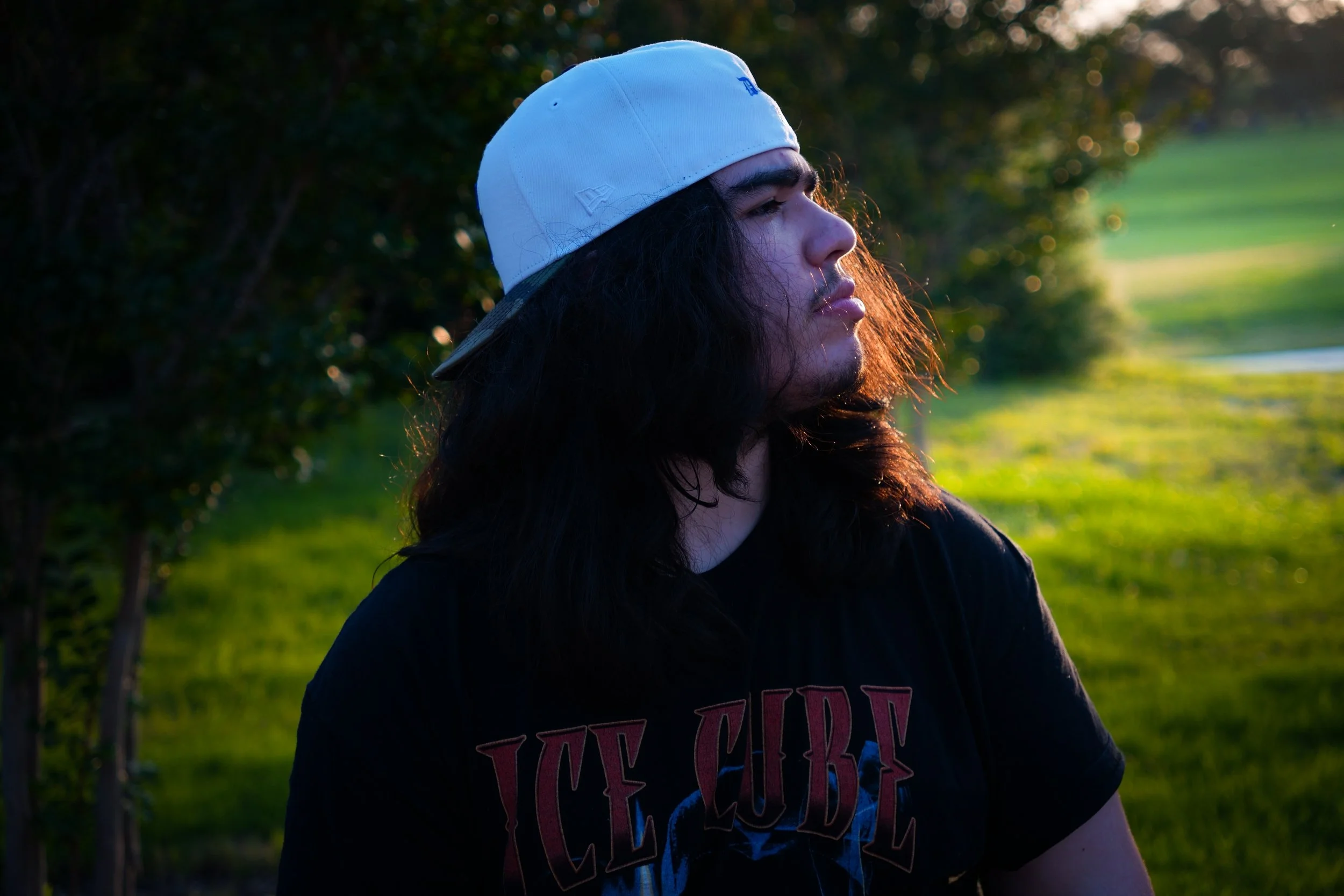 A young man with long dark hair wearing a white baseball cap backwards and an Ice Cube t-shirt, standing outdoors during sunset with greenery in the background.