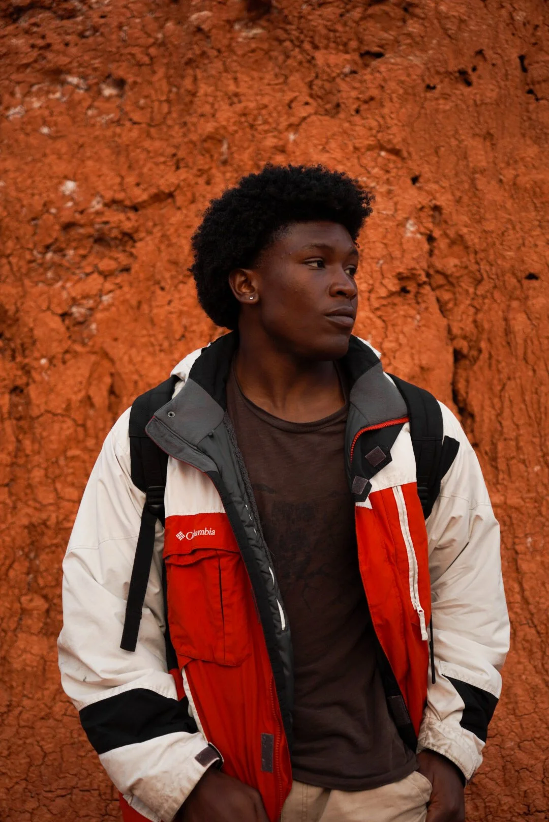A young man with curly black hair, wearing a Columbia jacket, standing against a textured red clay wall.