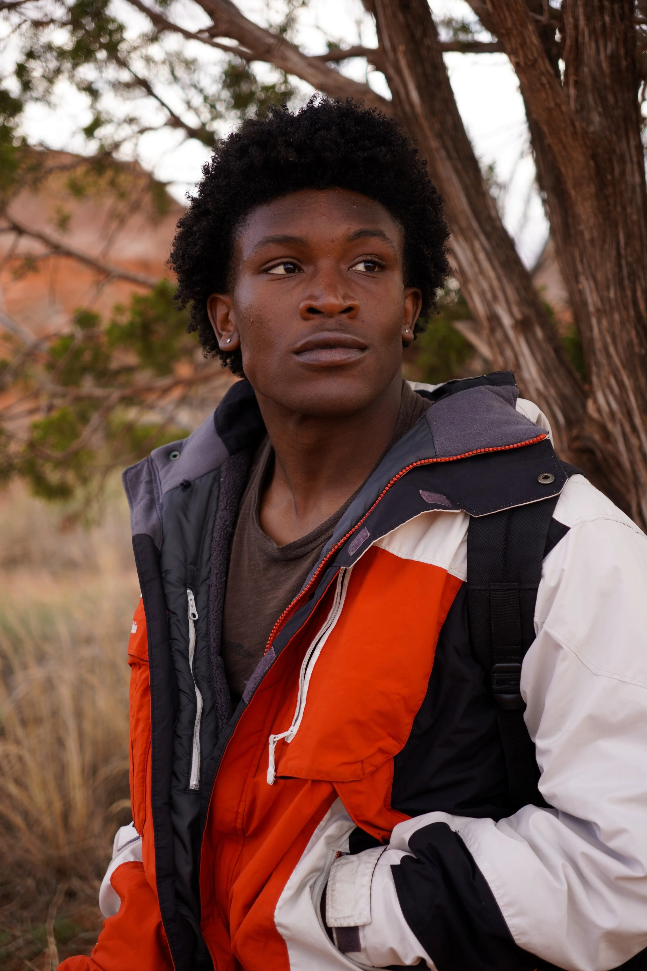 A young man with curly black hair, wearing earrings, a dark T-shirt, and a multicolored outdoor jacket with orange, black, white, and gray, stands outdoors near a tree with brown foliage and branches in the background.