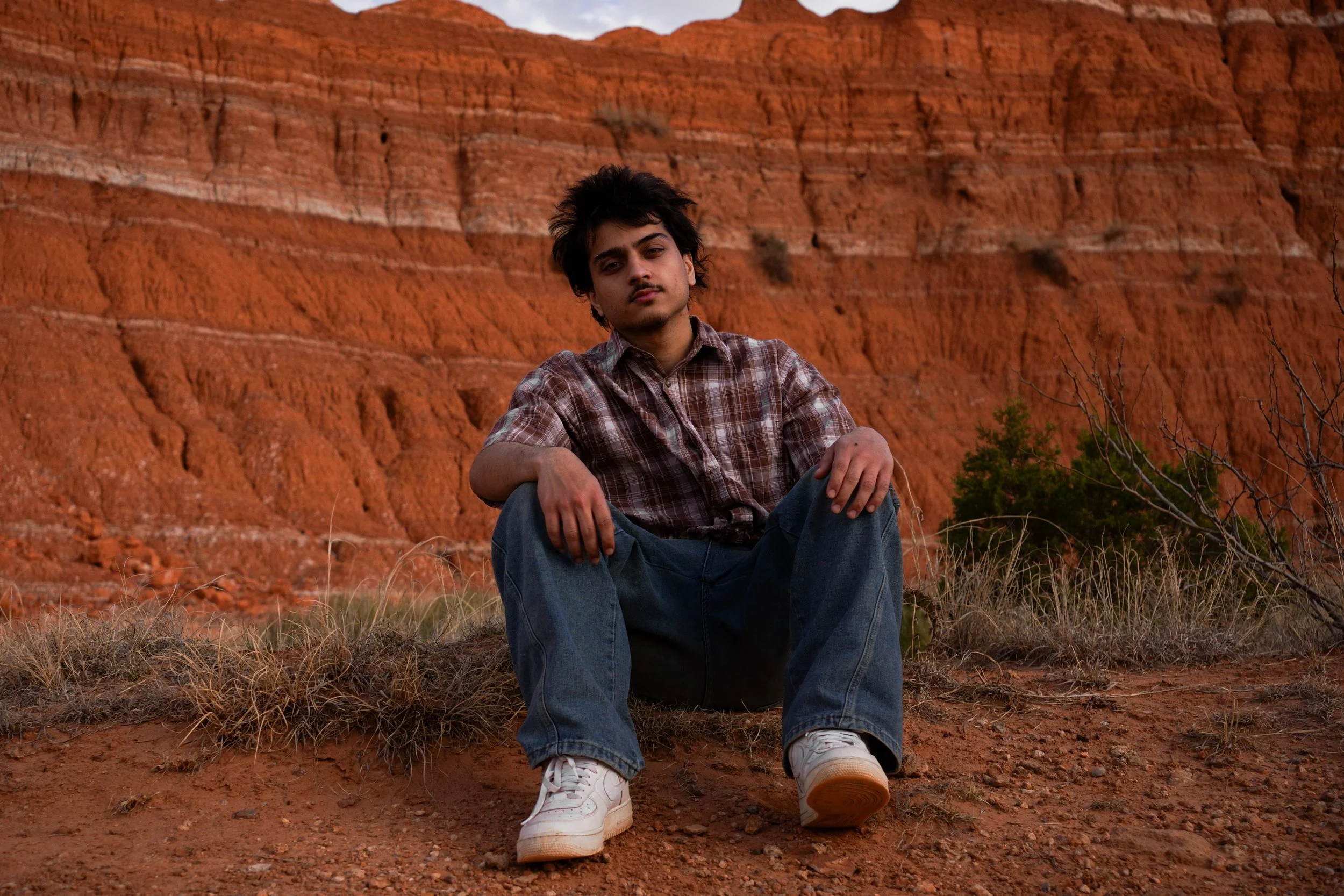 A young man with dark hair, wearing a plaid shirt, jeans, and white sneakers, sits on the ground in front of a red rock formation in a desert landscape.