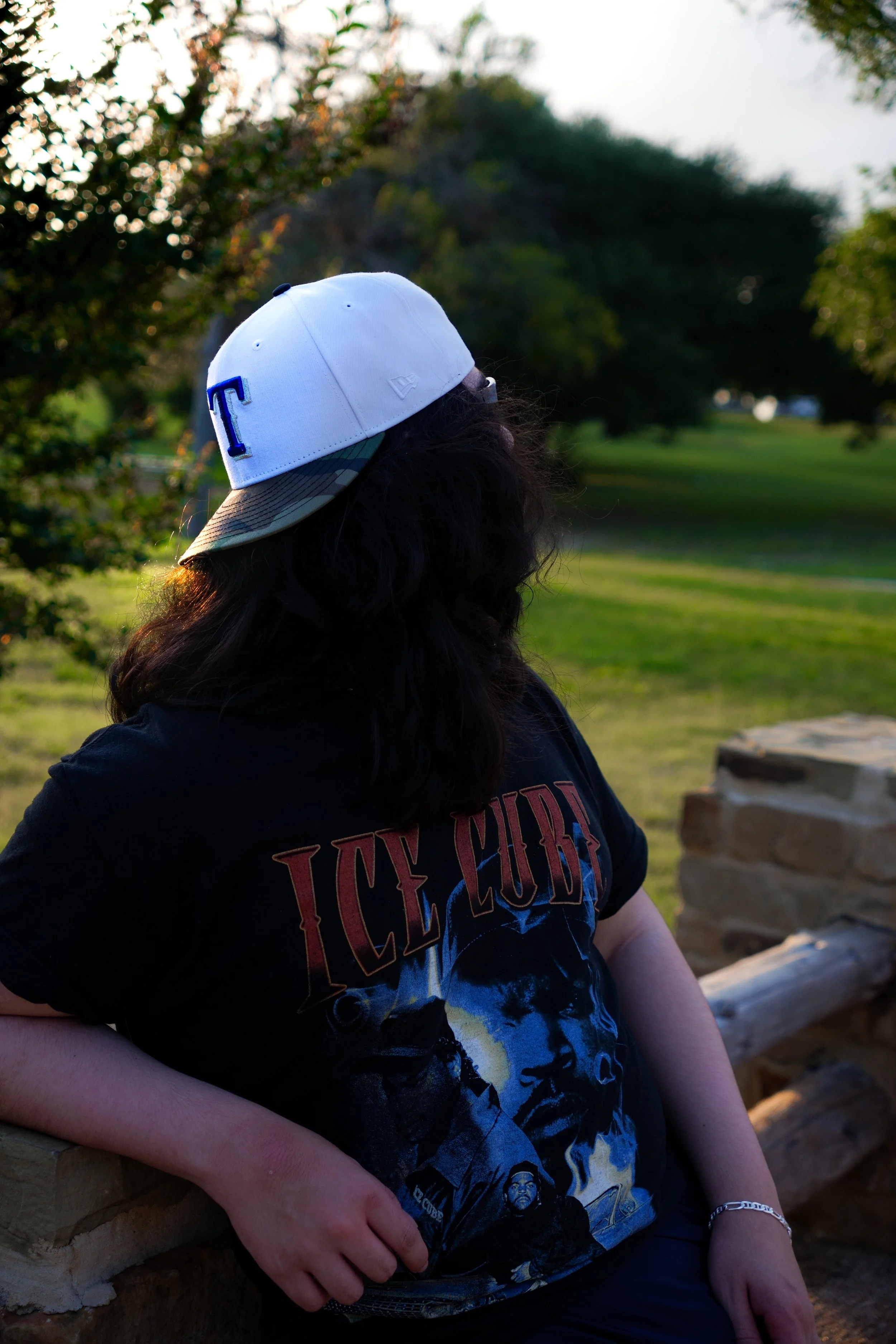 Person with curly dark hair wearing a white baseball cap backwards and a black Iced Cube band t-shirt, sitting outdoors on a stone ledge with trees and greenery in the background during sunset.