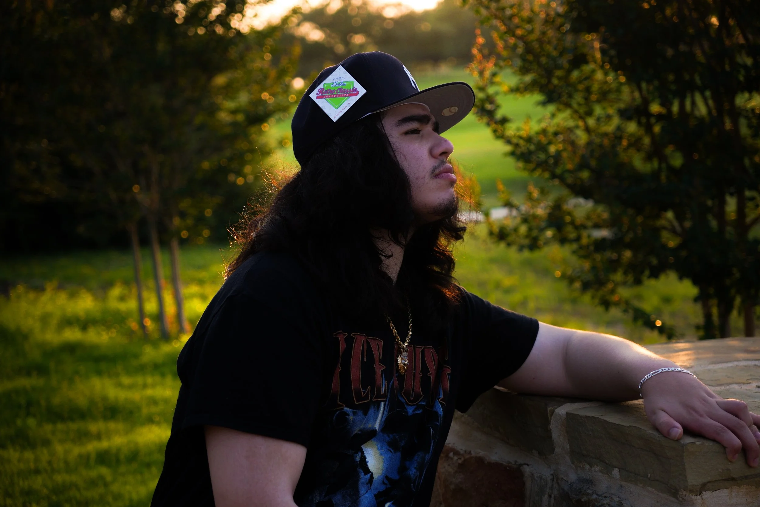 A young man with long dark hair, wearing a black baseball cap with a 'Reaga Classics' sticker, a gold chain, a silver bracelet, and a black Led Zeppelin T-shirt, leaning on a stone ledge outdoors during sunset with greenery and trees in the backgroun