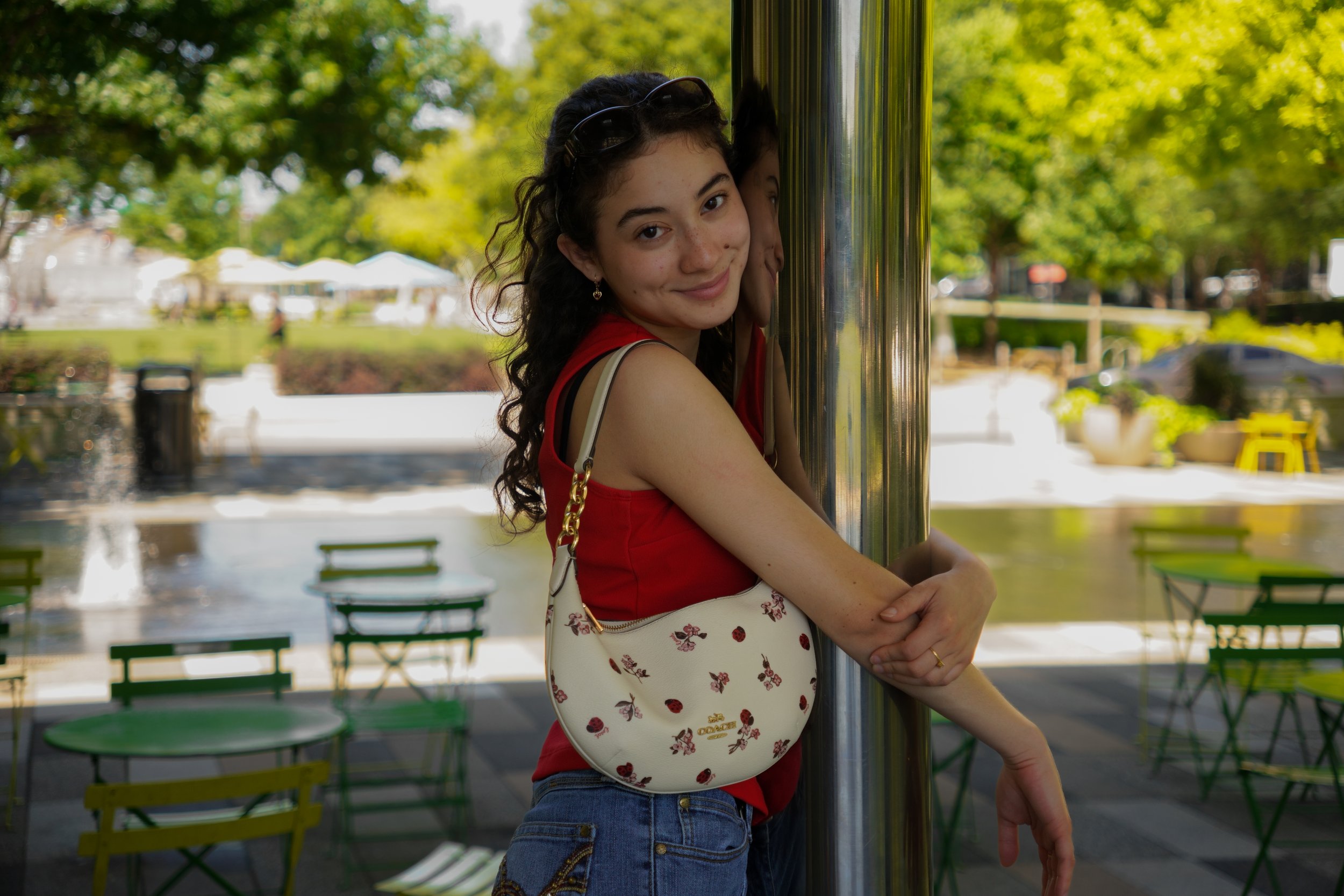 Young woman with long curly hair, wearing a red top and carrying a floral patterned purse, smiling and leaning against a metallic pole outdoors in a park with trees and empty tables.