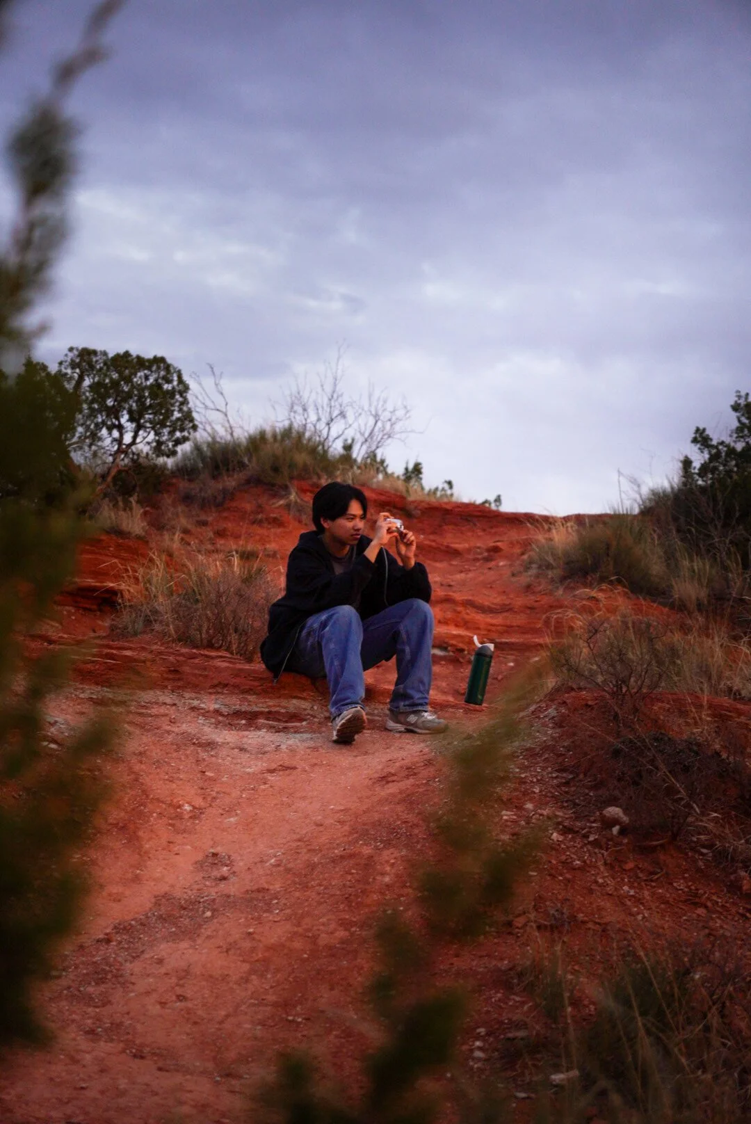 Young man sitting on a dirt trail in a desert landscape, taking a photo with a camera, surrounded by reddish soil, bushes, and sparse vegetation, under a cloudy sky.