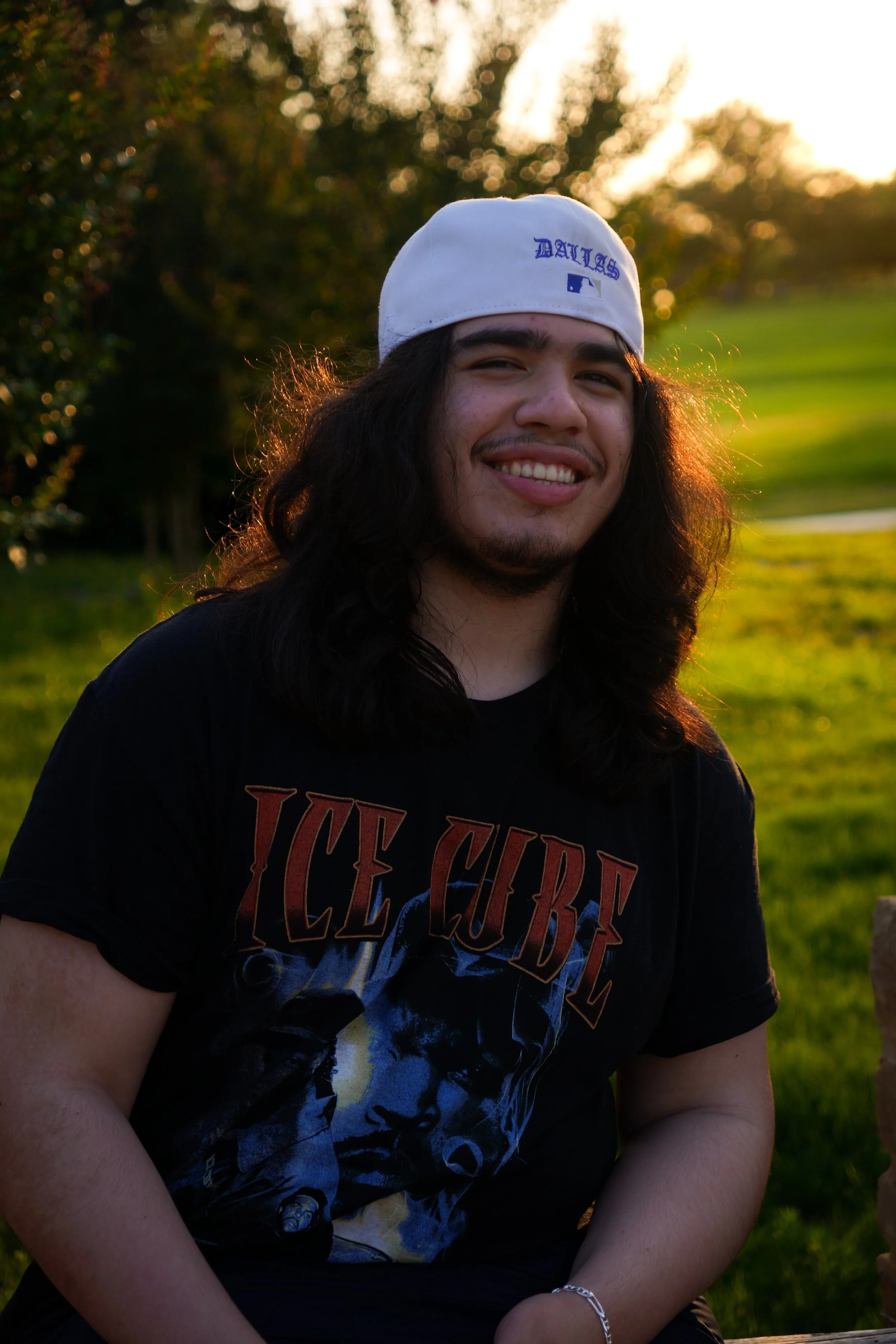 A young man with long dark hair, smiling, sitting outdoors during sunset. He is wearing a white cap with blue text and a black t-shirt featuring an Ice Cube graphic.