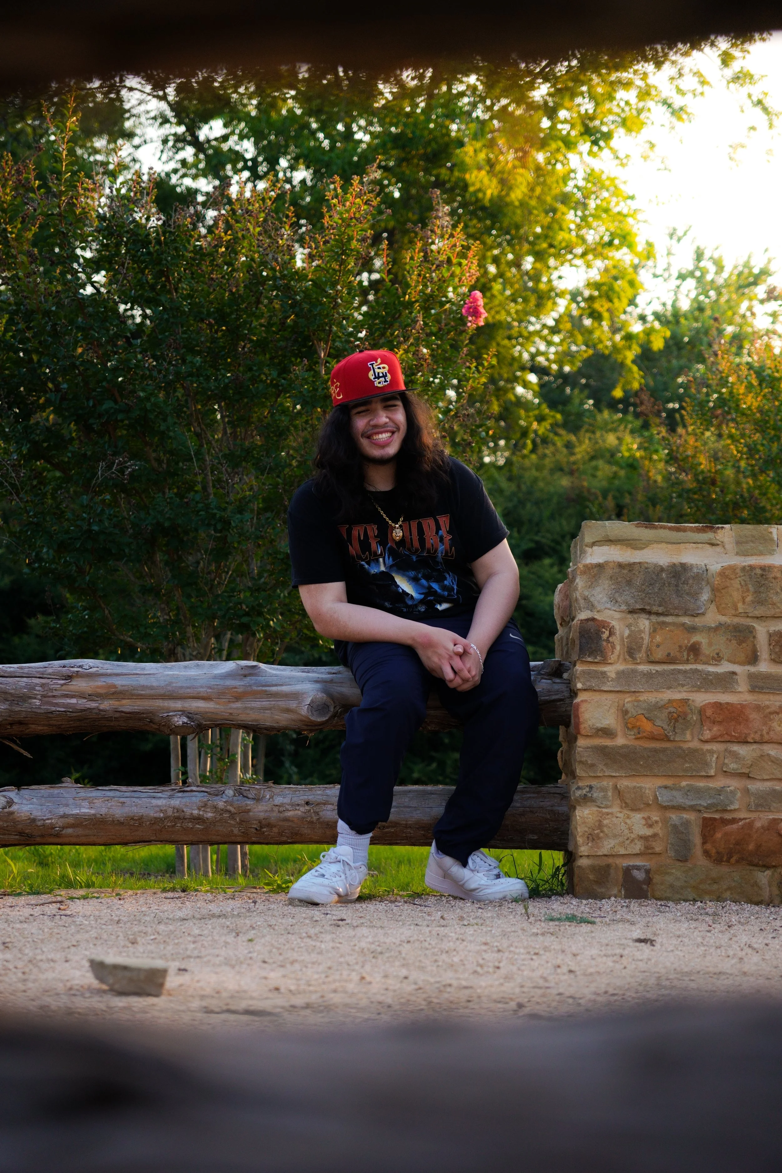 Young man sitting on a log outdoors, smiling, wearing a black T-shirt, navy pants, white sneakers, and a red cap, with greenery and a stone wall in the background.