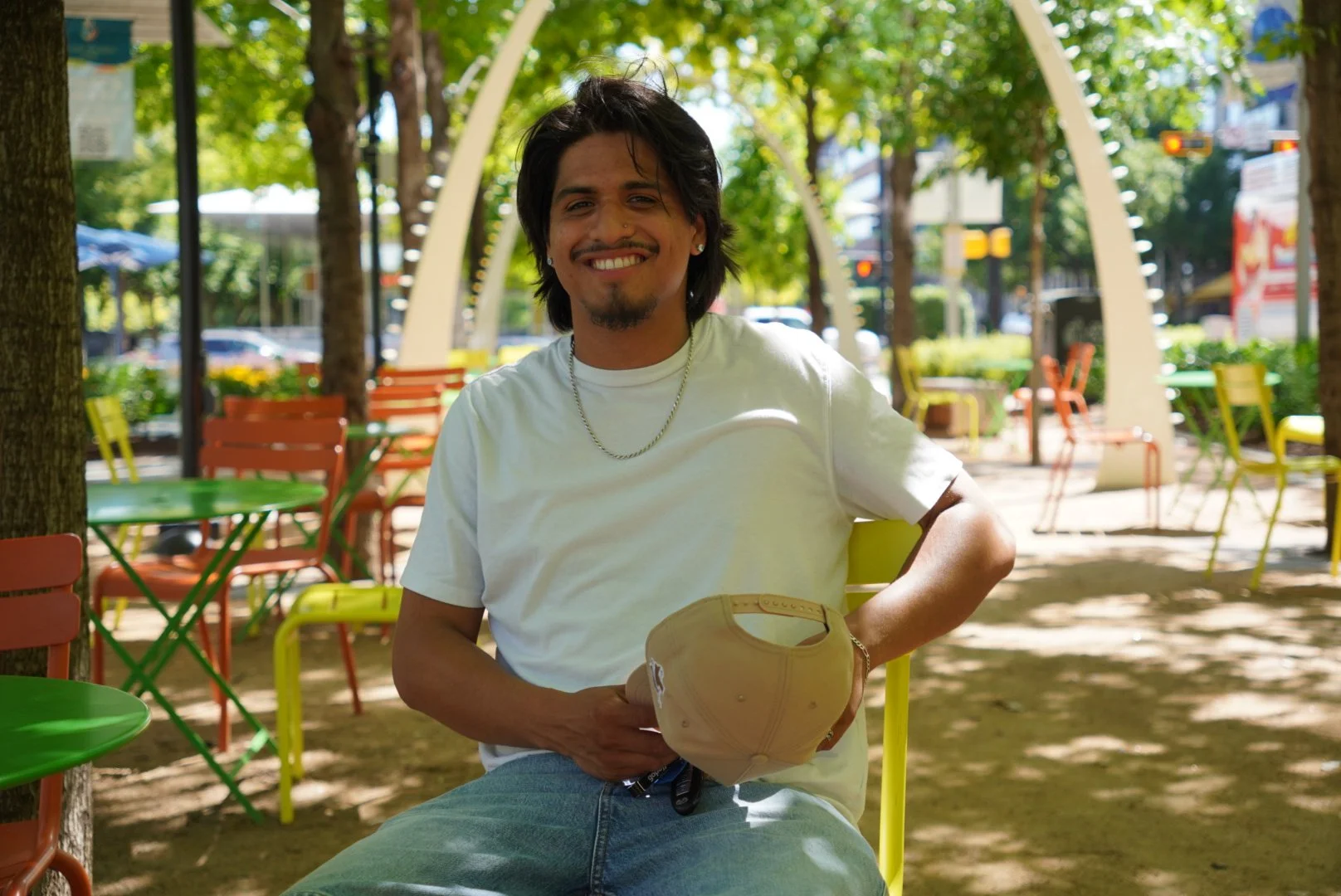 A young man with dark hair and a goatee smiling and sitting at an outdoor cafe with colorful chairs and trees in the background, holding a beige baseball cap.