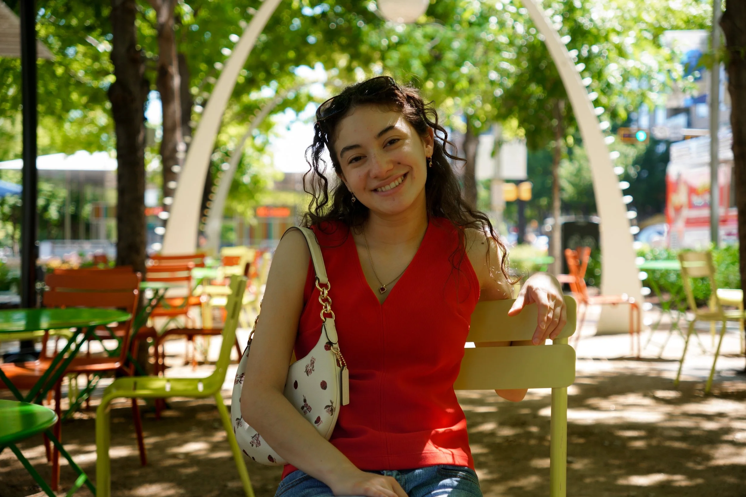 A young woman with curly dark hair smiling while sitting on a yellow chair outdoors, surrounded by colorful chairs and green trees, with a bright, sunny day.