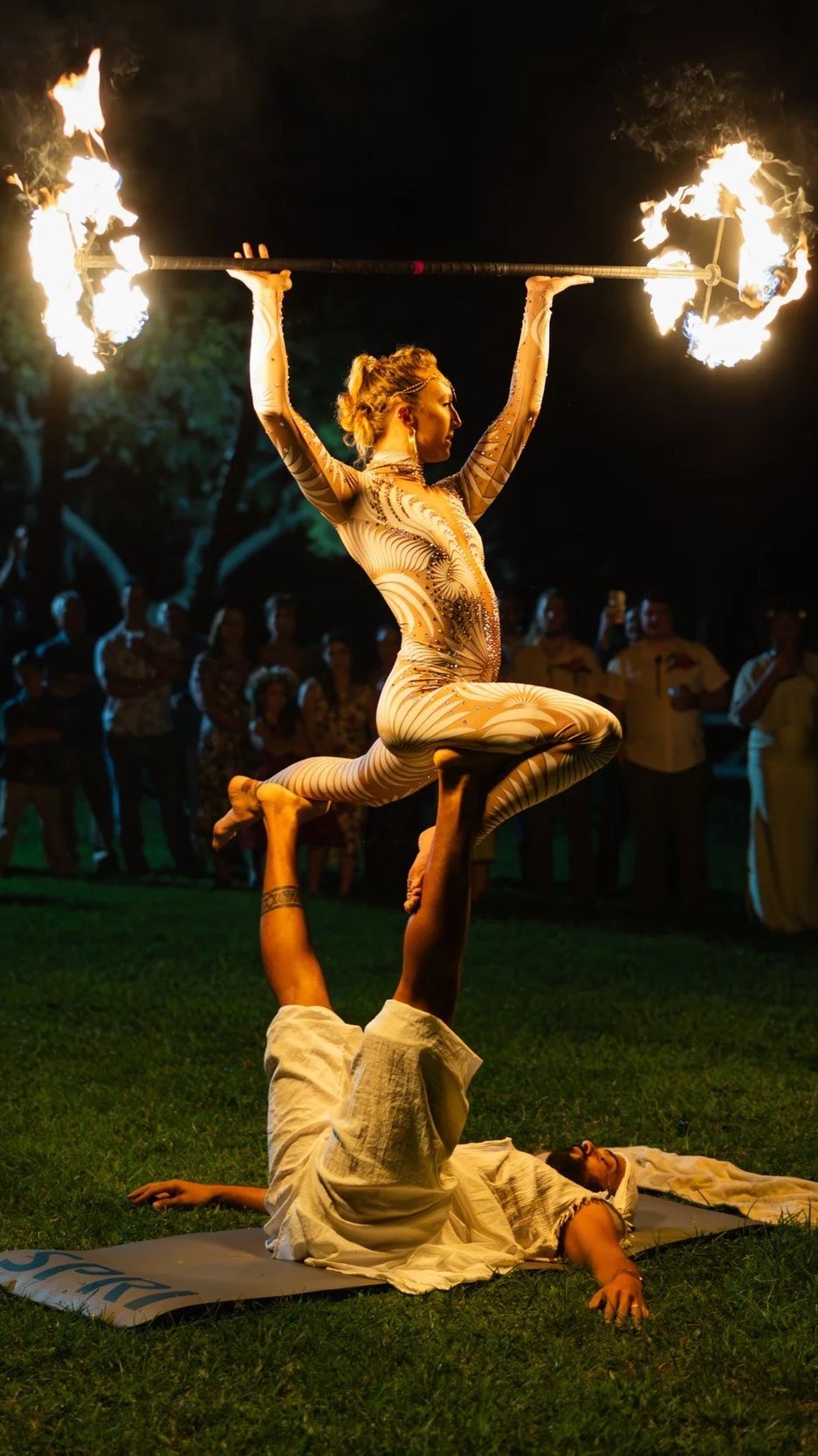 A female fire dancer in an intricate costume performs a balancing act with fire props, supported by a male performer lying on the ground, at night with a crowd watching.