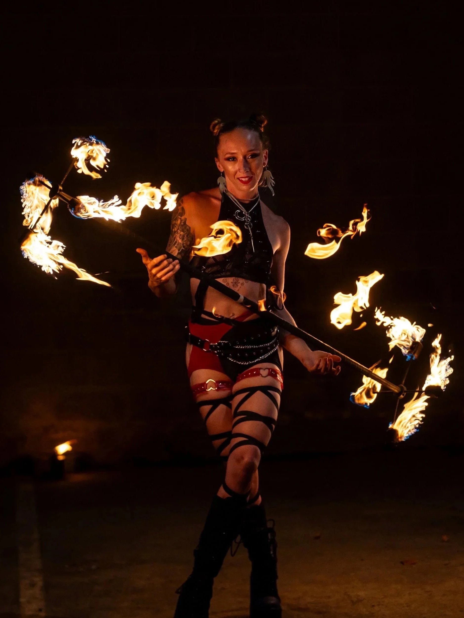 A woman performing fire spinning outdoors at night, holding a staff with flames on either end, wearing black and red clothing with tattoos and large earrings.
