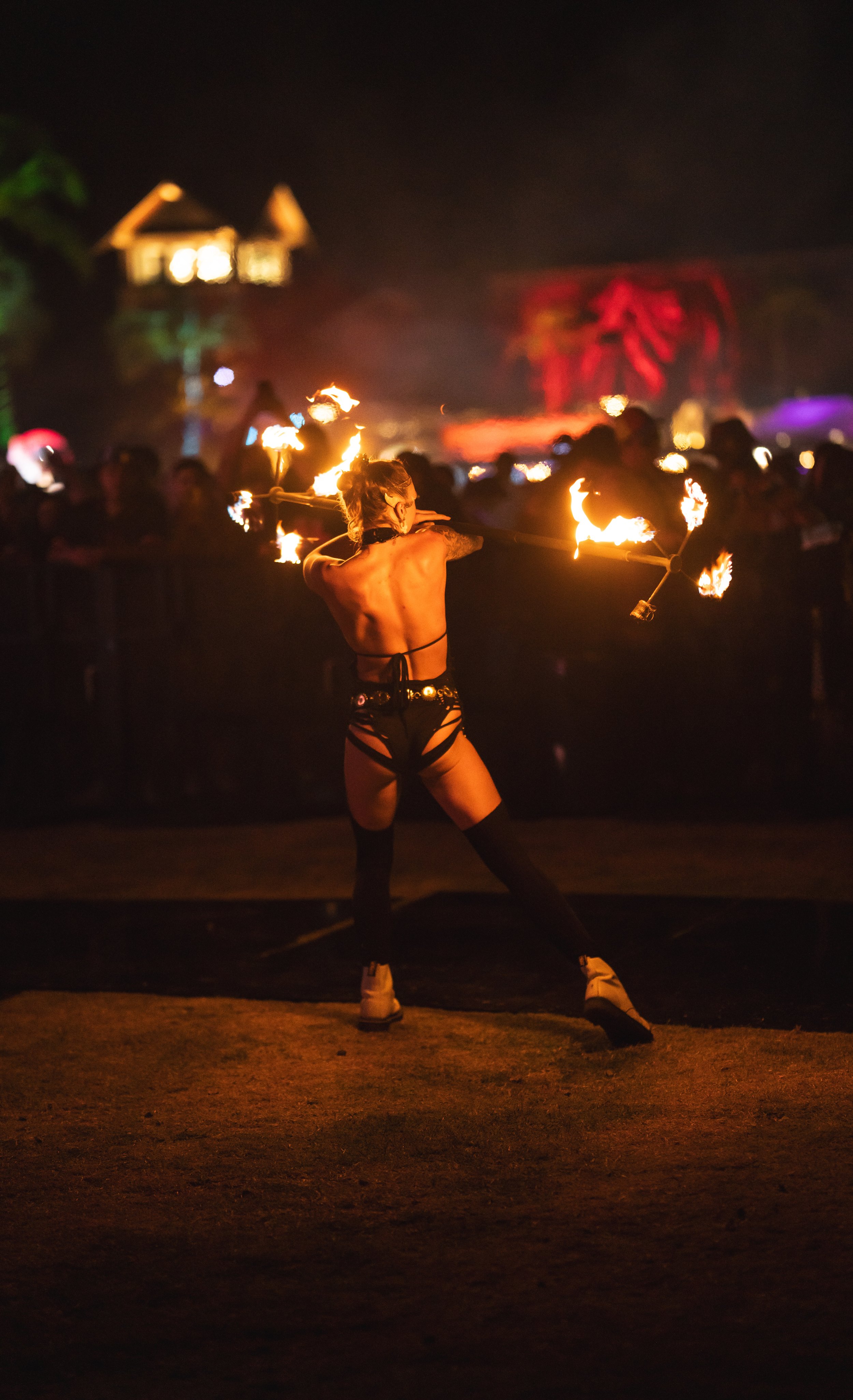 A fire performer with a bare back, wearing black leather straps and boots, spinning fire poi during a night event with blurred onlookers and colorful lights in the background.