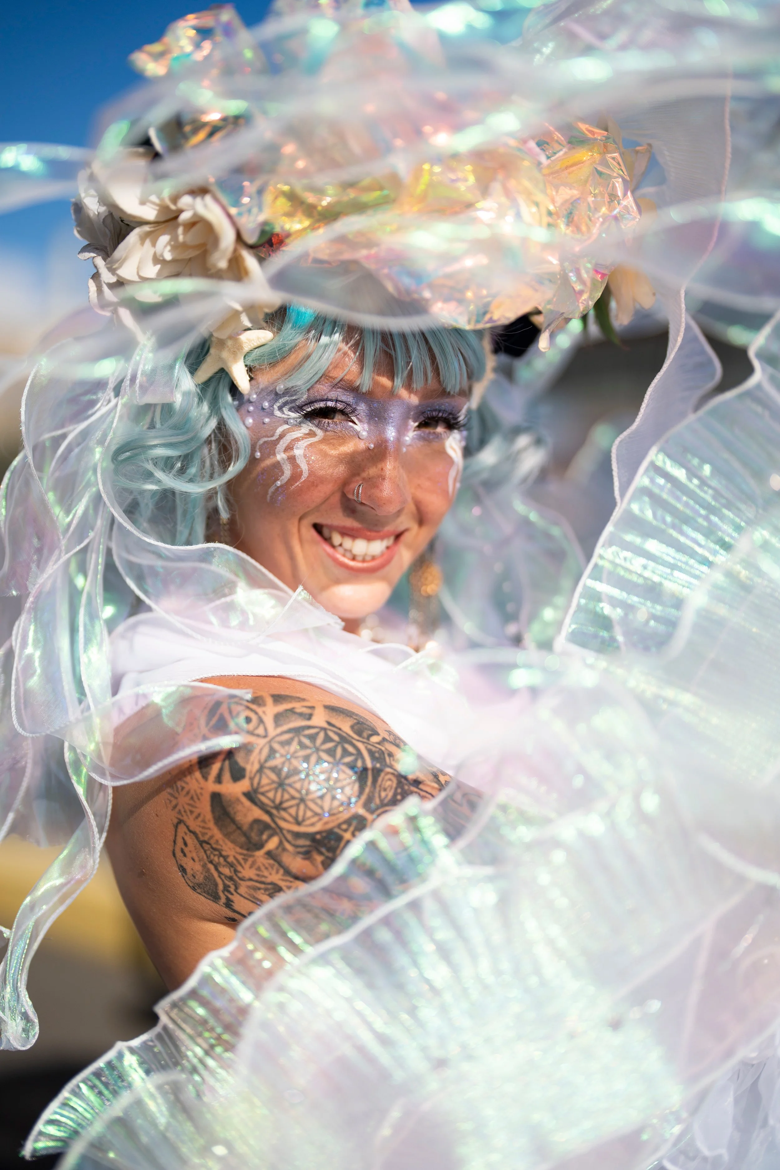 A person smiling with light blue hair, elaborate makeup, and a colorful costume adorned with iridescent and translucent materials, including seashells and starfish, on their head and shoulder, set against a bright outdoor background.