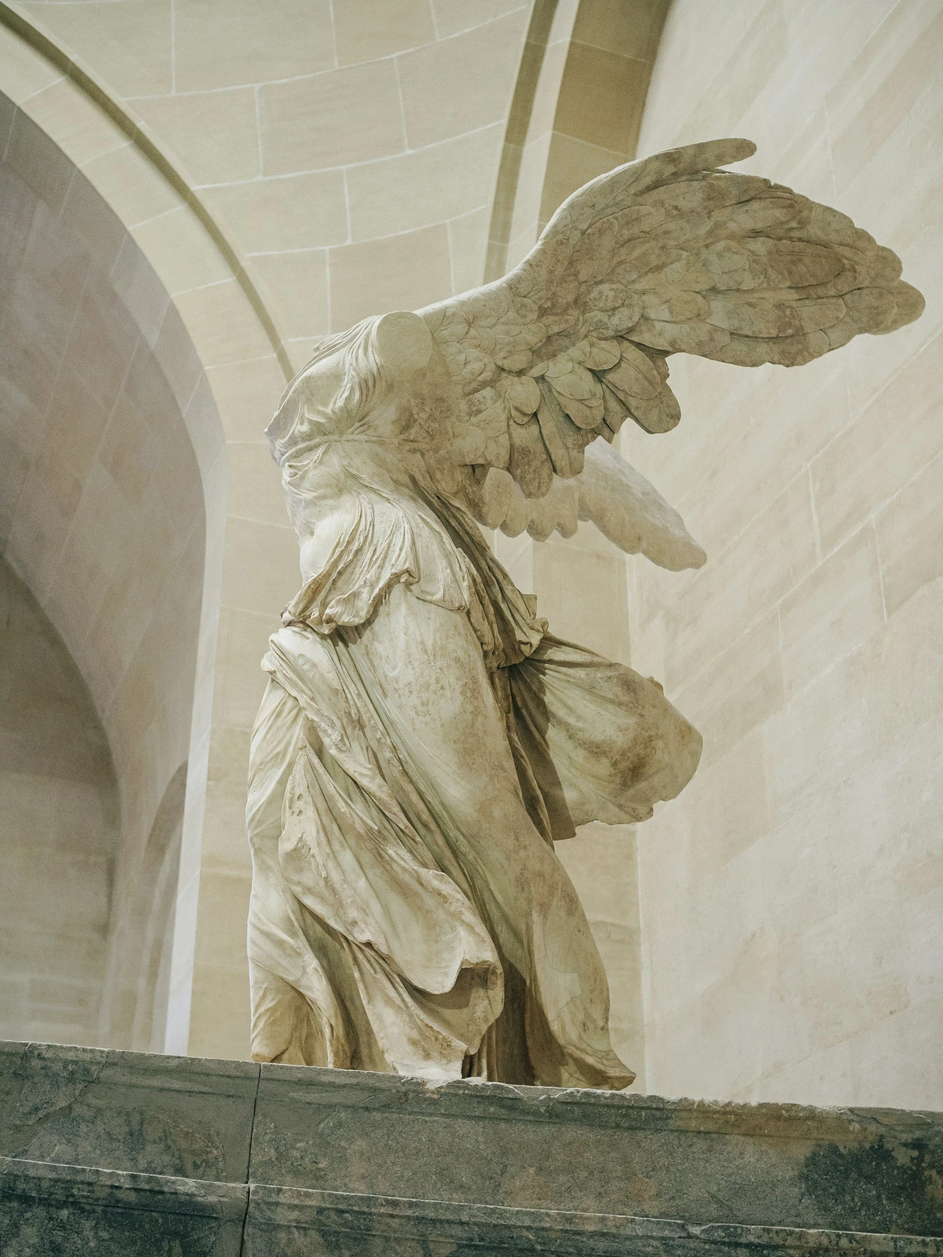 Marble statue of a winged figure, likely Nike, the Greek goddess of victory, displayed in a museum or temple setting with a stone arch in the background.