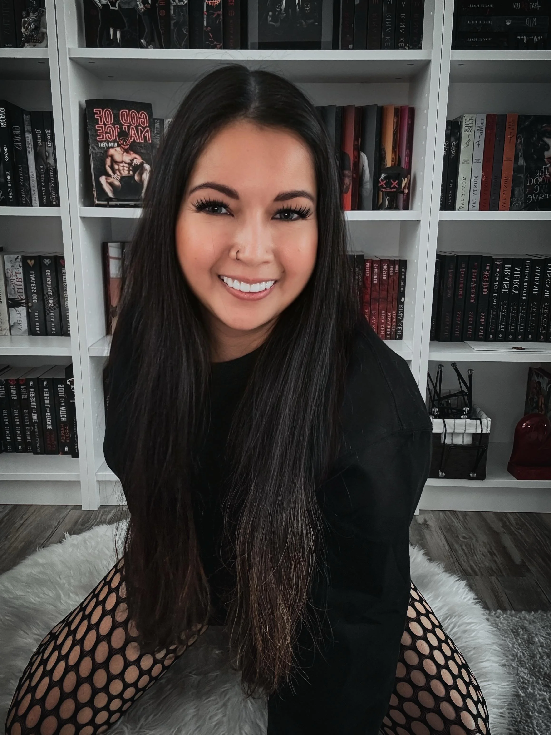 PR Team Member Lala, posed on the floor, in front of a bookshelf. Wearing a black shirt, with long brown hair.