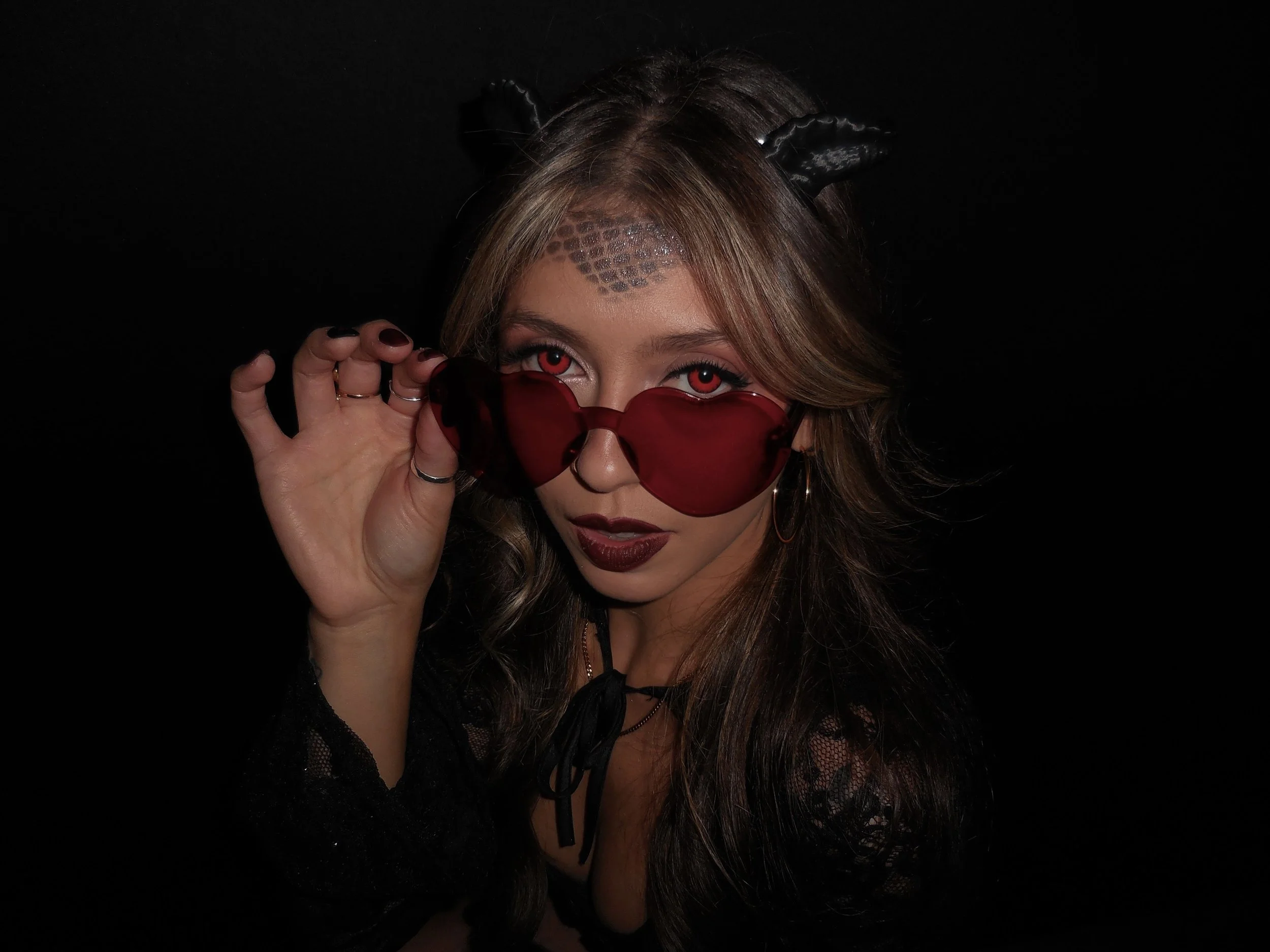 A woman with wavy, shoulder-length hair wearing dark lipstick, layered necklaces, and a black strapless corset dress, against a black background.