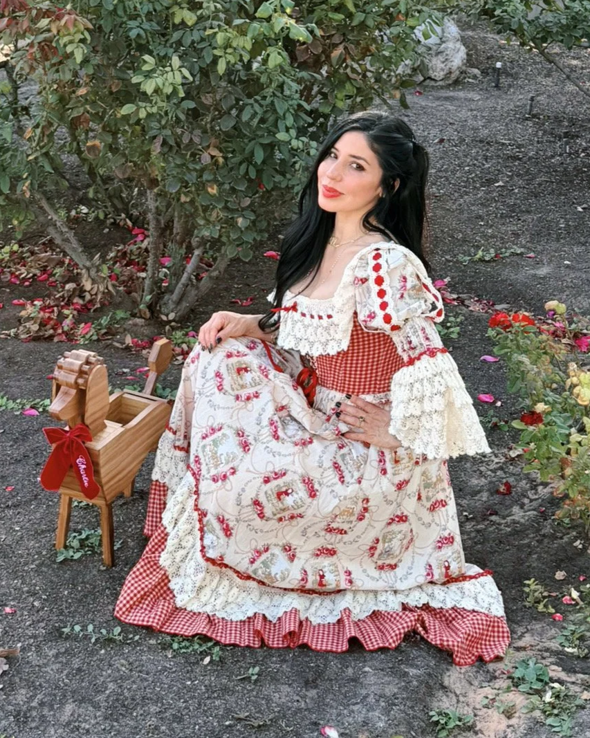 A woman dressed in a vintage, country-style dress with red and white patterns, sitting on the ground near a hedge with red and pink flowers. She has long black hair, red lipstick, and is smiling slightly. A small wooden rocking horse with a red bow is beside her.