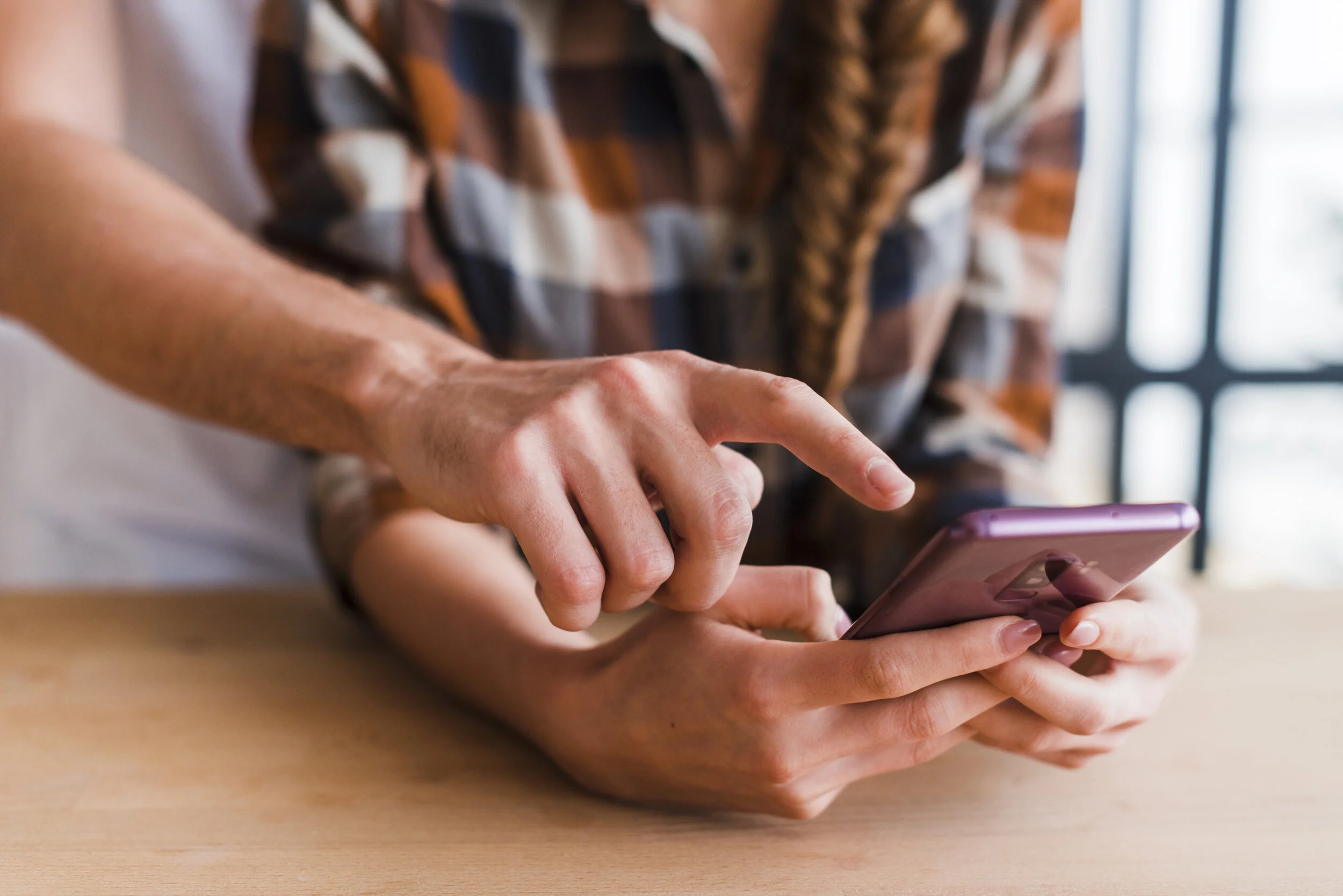 Close-up of two people holding smartphones, one person using a pink phone with a purple grip and the other person's hand is seen reaching for it, with a window in the background.