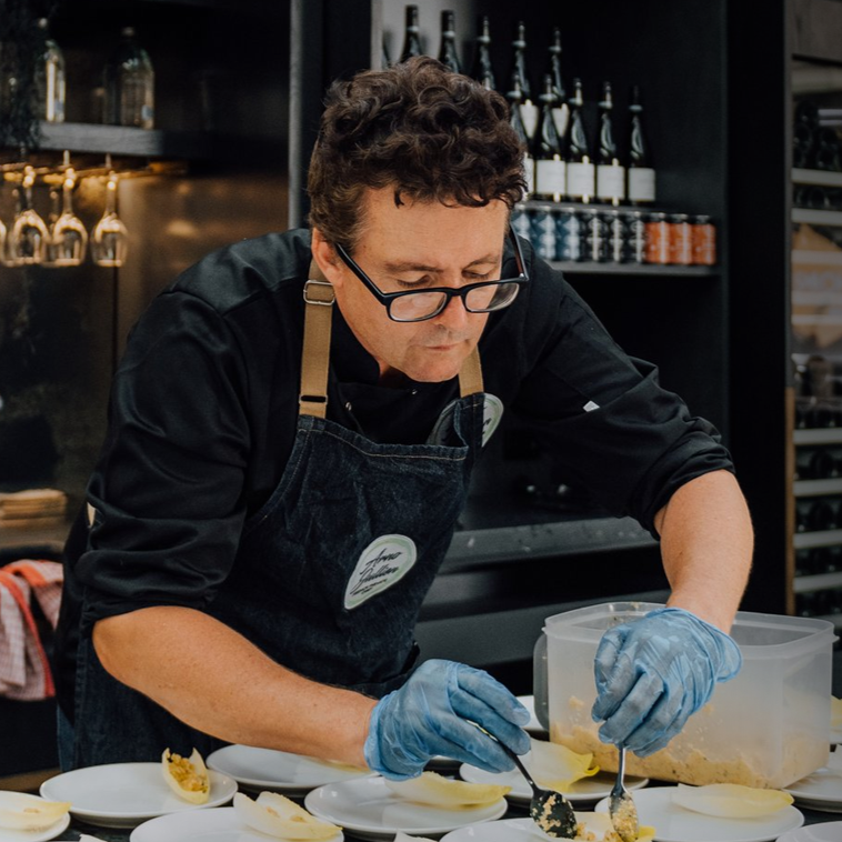 A man wearing a black chef jacket with a black shirt, apron, and blue gloves is preparing food in a modern kitchen or food preparation area, filling white bowls with a yellow filling.
