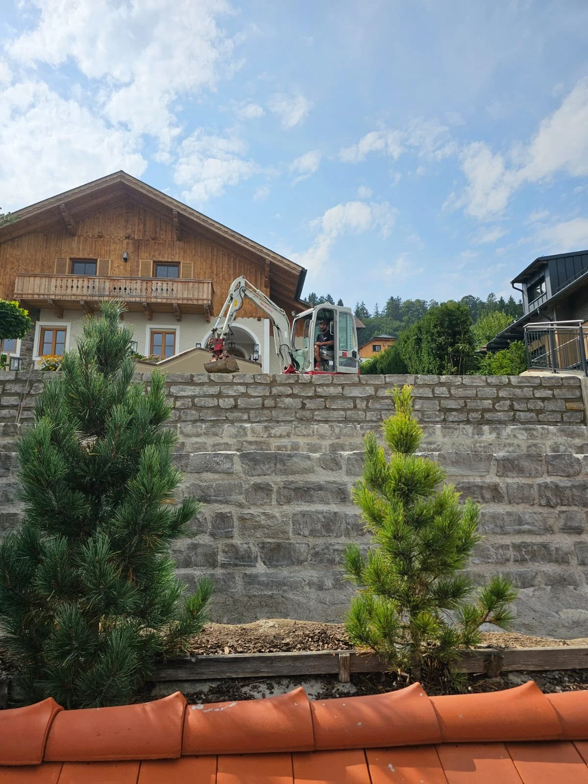 Ein Bauarbeiter fährt mit einem kleinen Bagger auf einer Steinmauer in einem Wohngebiet mit grünen Bäumen und mehreren Häusern im Hintergrund, bei sonnigem Himmel.