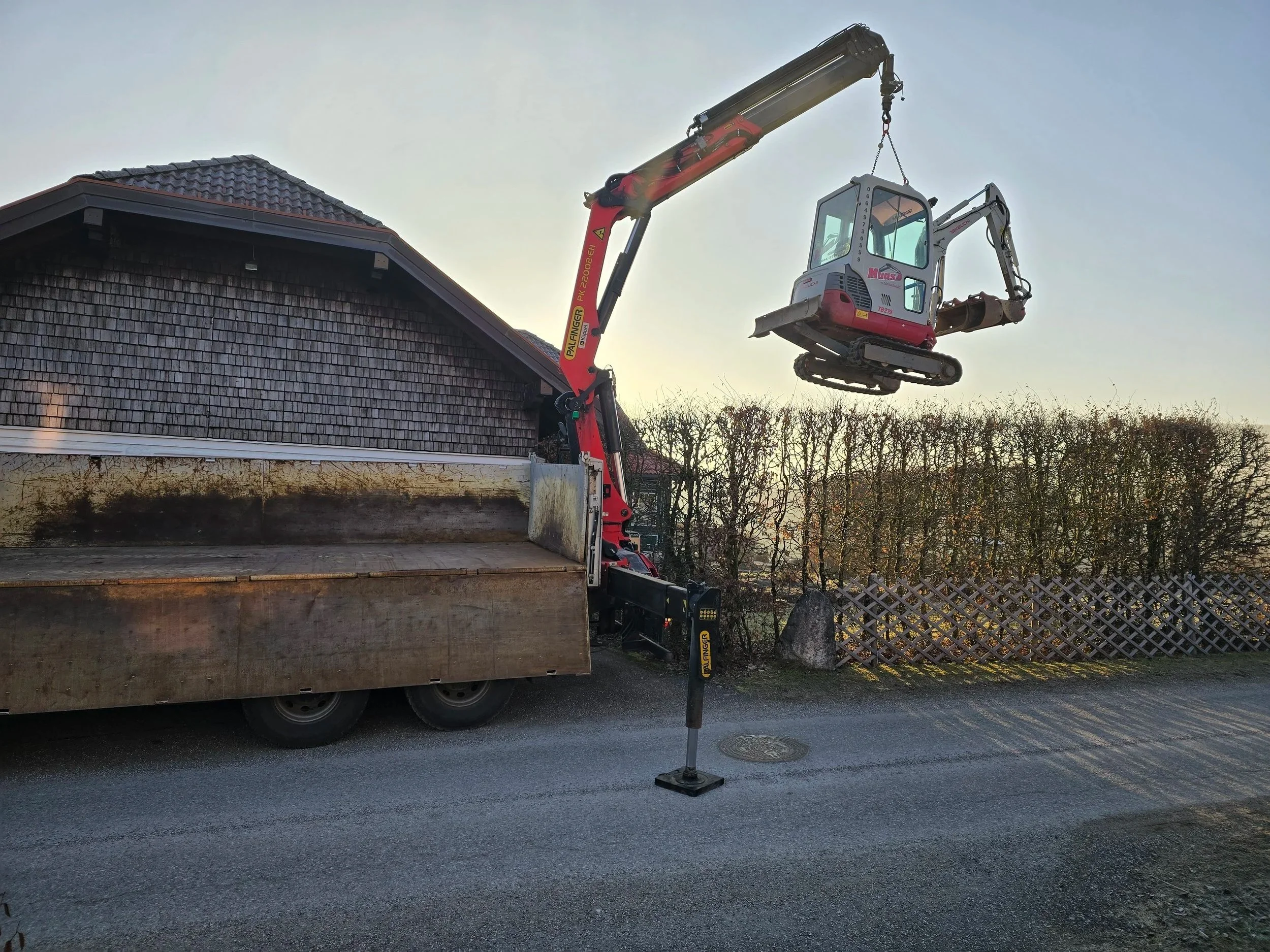 Ein Baukran hebt eine kleine Baumaschine in die Luft neben einem Haus mit dunklem Ziegeldach. Der Baukran befindet sich auf einer Straße, und im Hintergrund sind Bäume und ein Holzzaun sichtbar.
