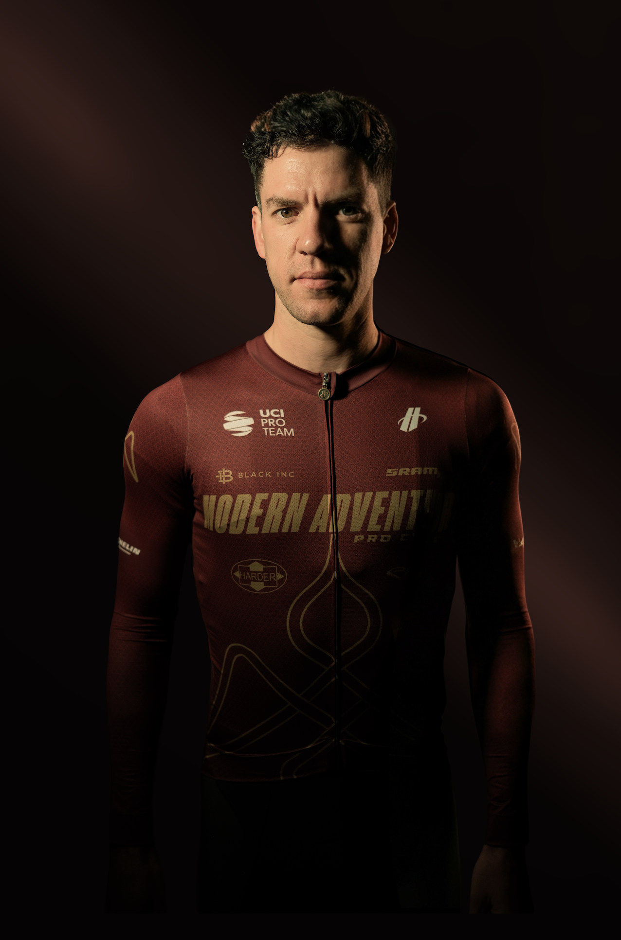 Portrait of a young man in a dark red cycling jersey with various sponsor logos, standing against a dark background.