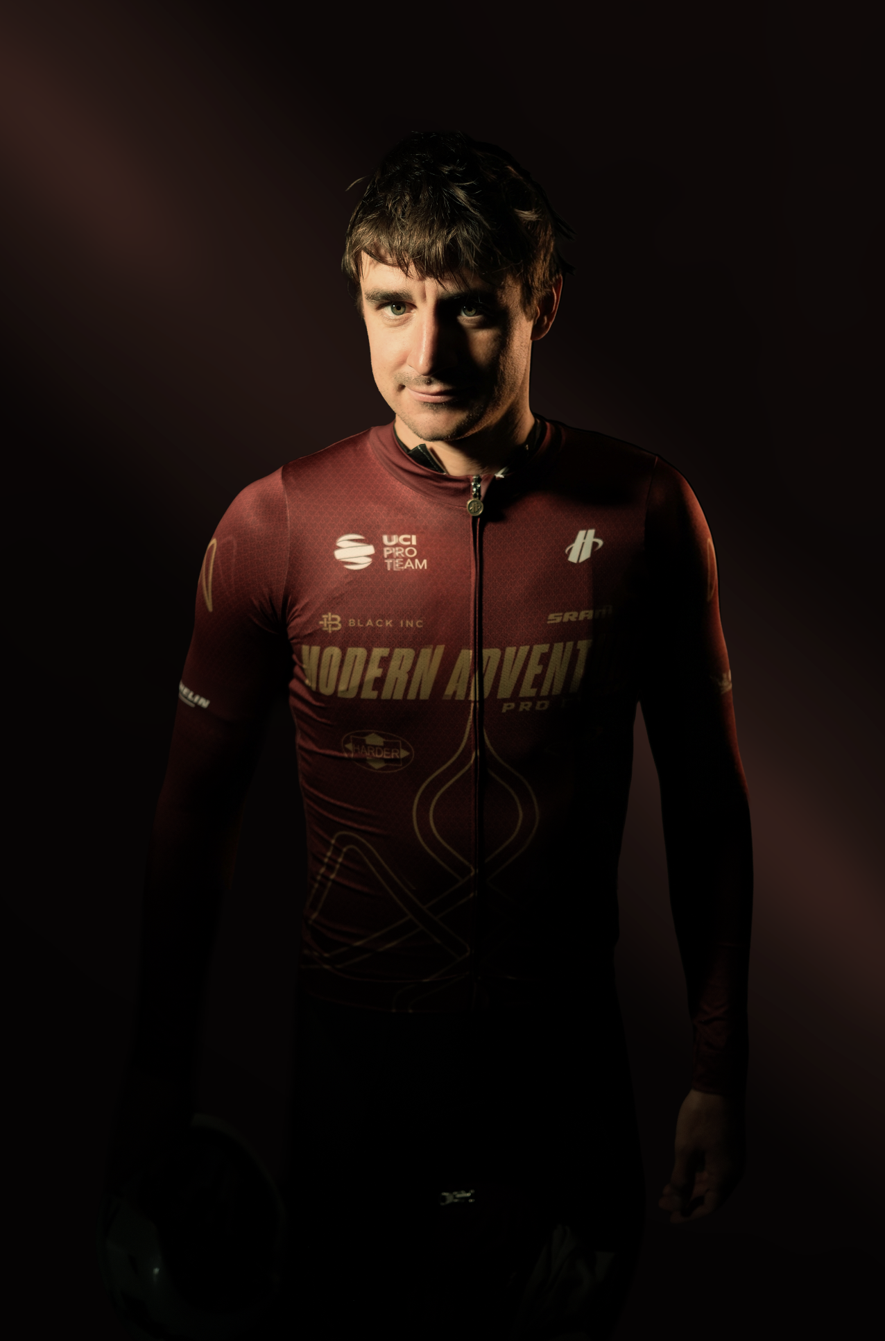 A male cyclist in a dark red riding uniform standing with a slight smile, holding a helmet, against a dark background.