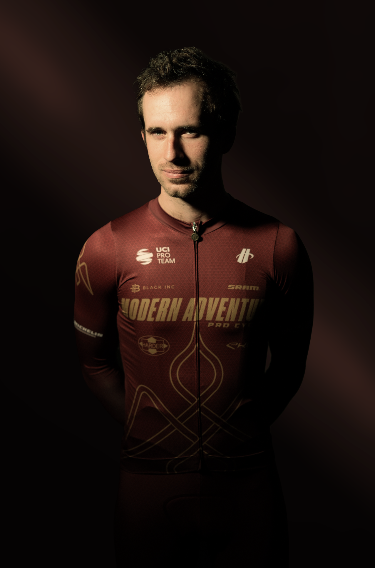 A man in a red cycling jersey with gold accents and various sponsor logos, standing against a dark background.