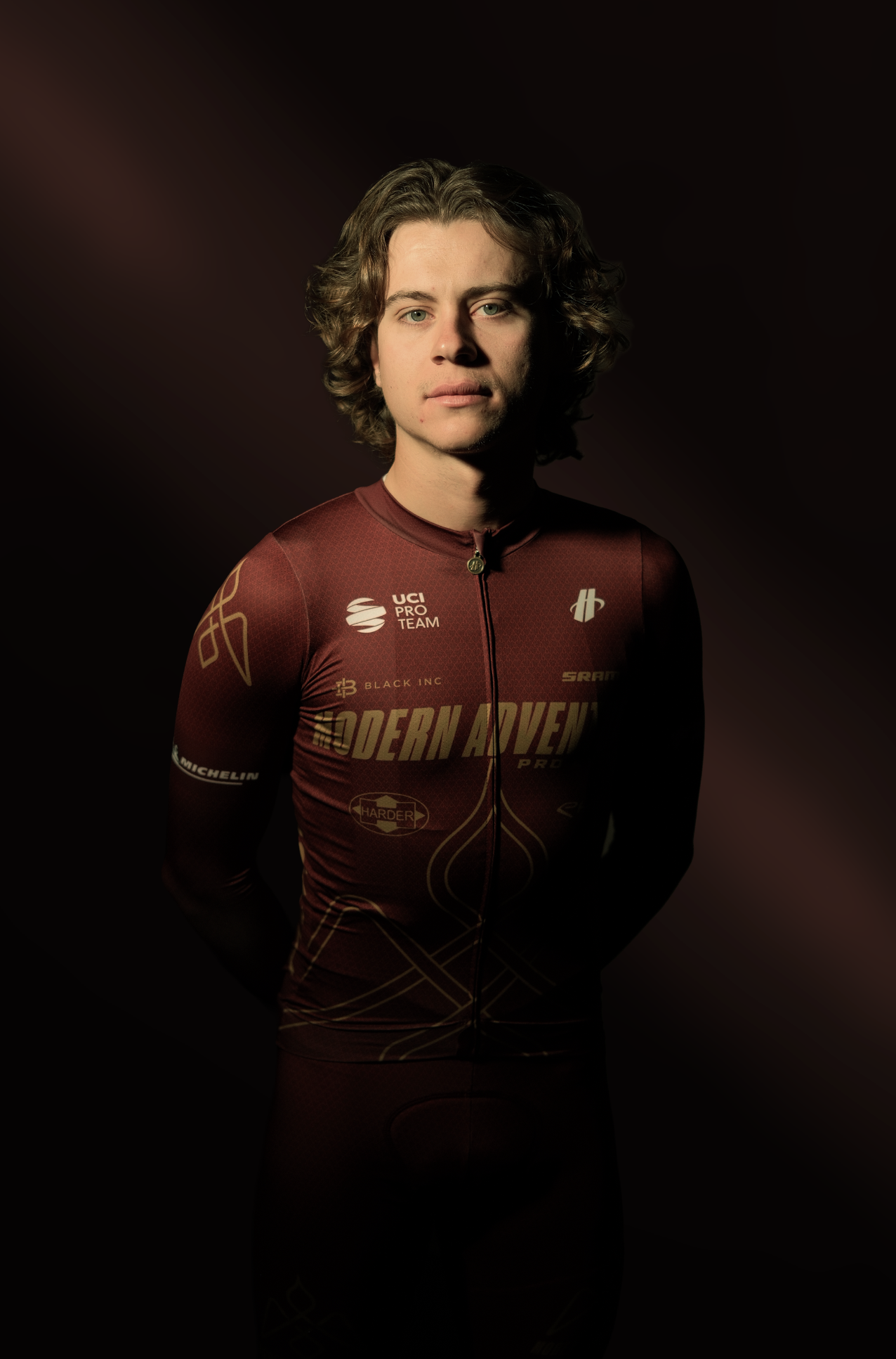 Young male cyclist with wavy brown hair, wearing a maroon cycling jersey with gold and white graphics and sponsors, standing against a dark background.