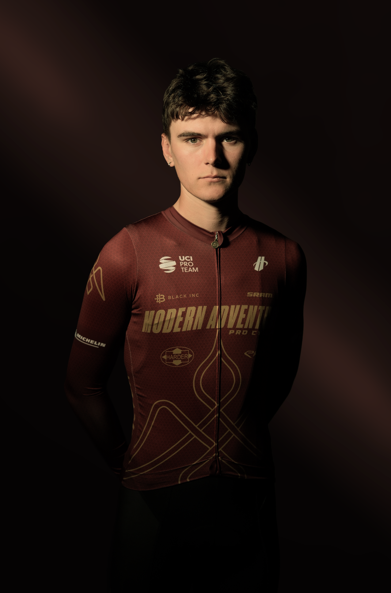 Young woman in a maroon cycling jersey with various logos and patterns, standing against a dark background.