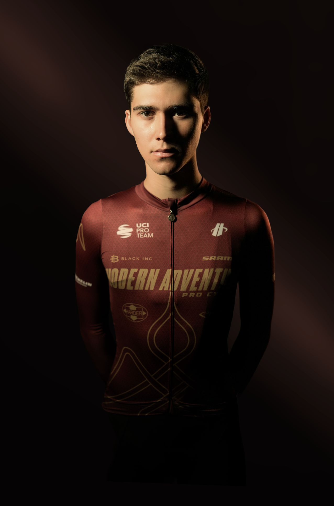 A young man wearing a maroon cycling jersey with various sponsorship logos, standing against a dark background.