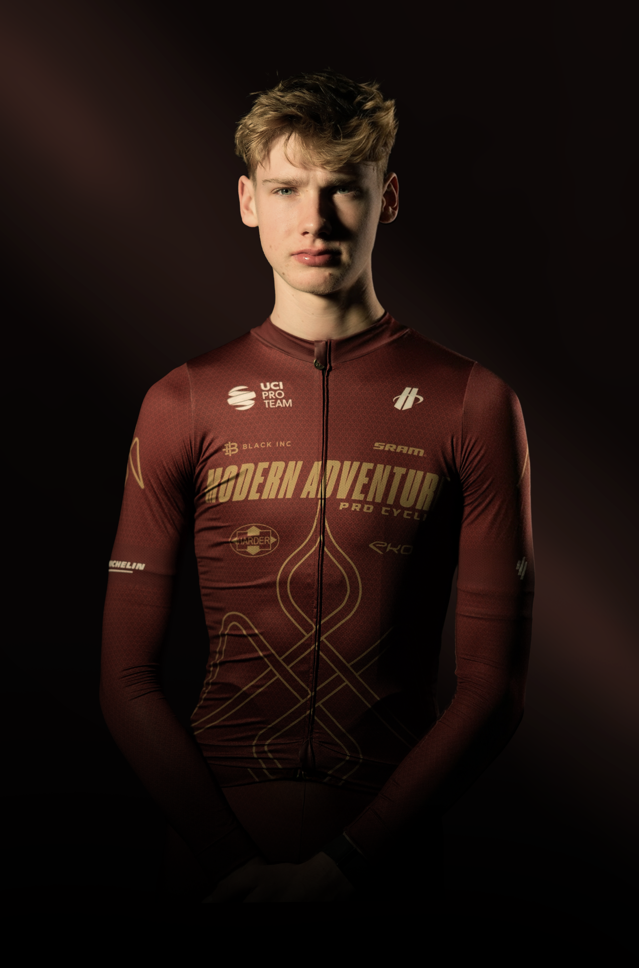 Young man wearing a maroon cycling jersey with various logos, standing against a dark background.