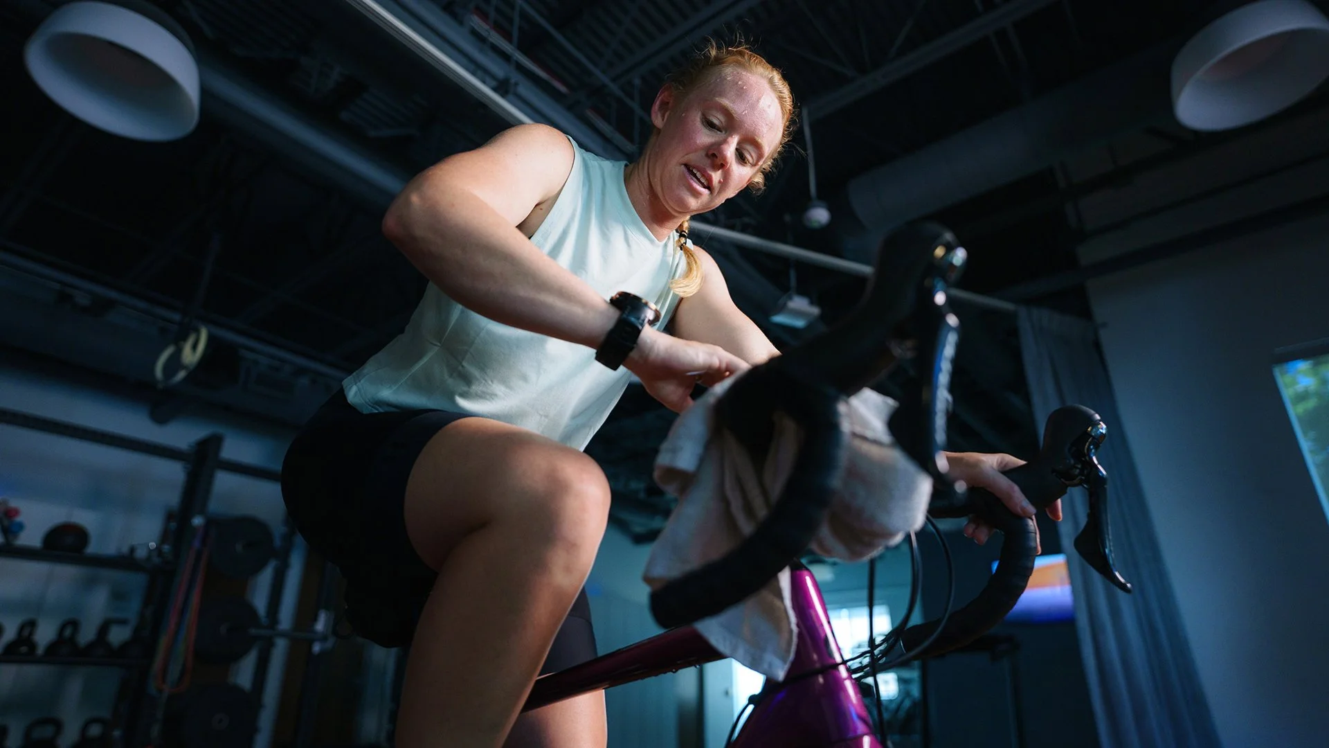 A woman with red hair and fair skin riding a stationary exercise bike in a gym, looking focused.
