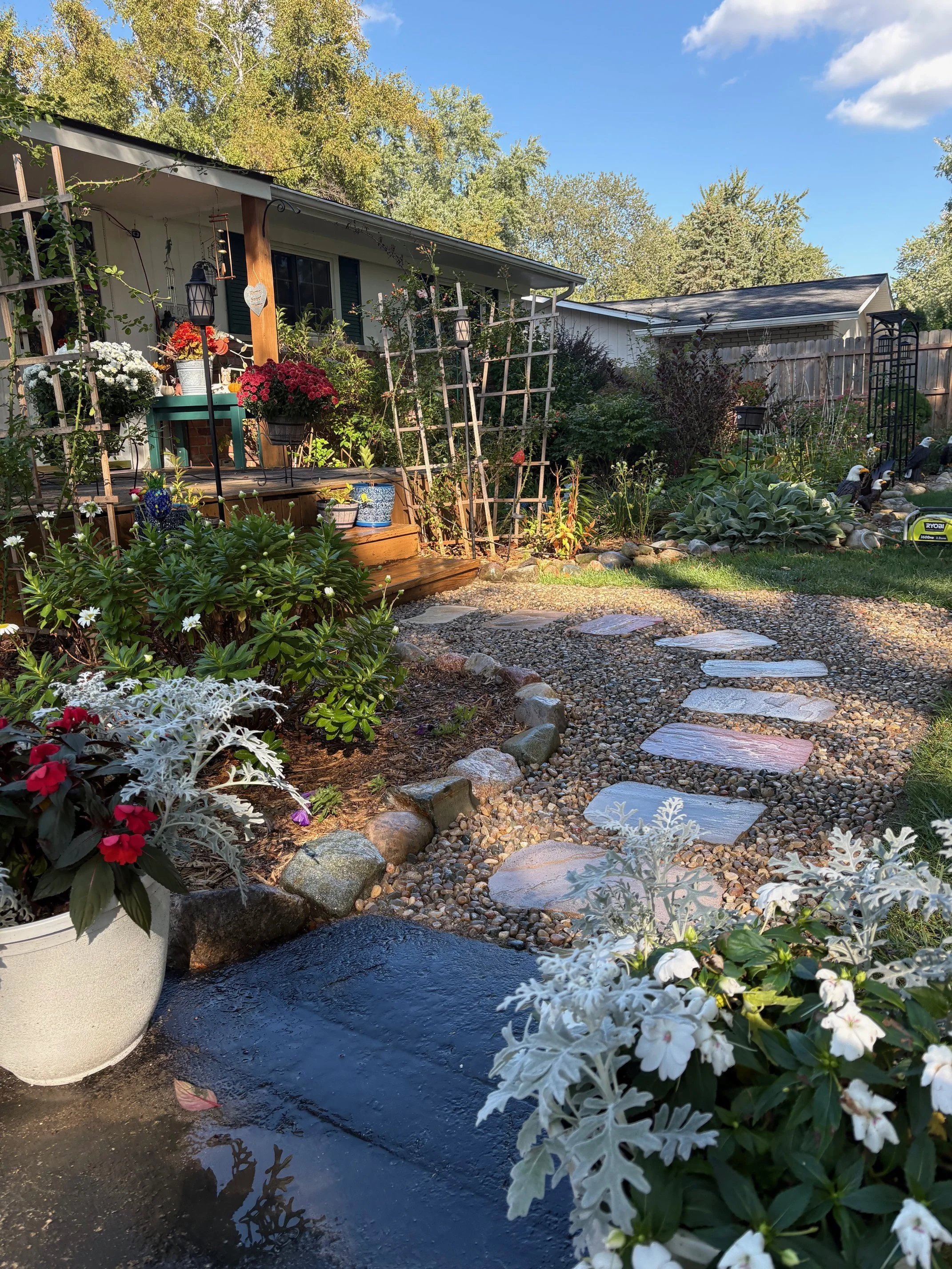 A backyard garden with a stepping stone pathway, flower pots, lush greenery, and a wooden deck with outdoor decorations and potted plants, under a clear blue sky.