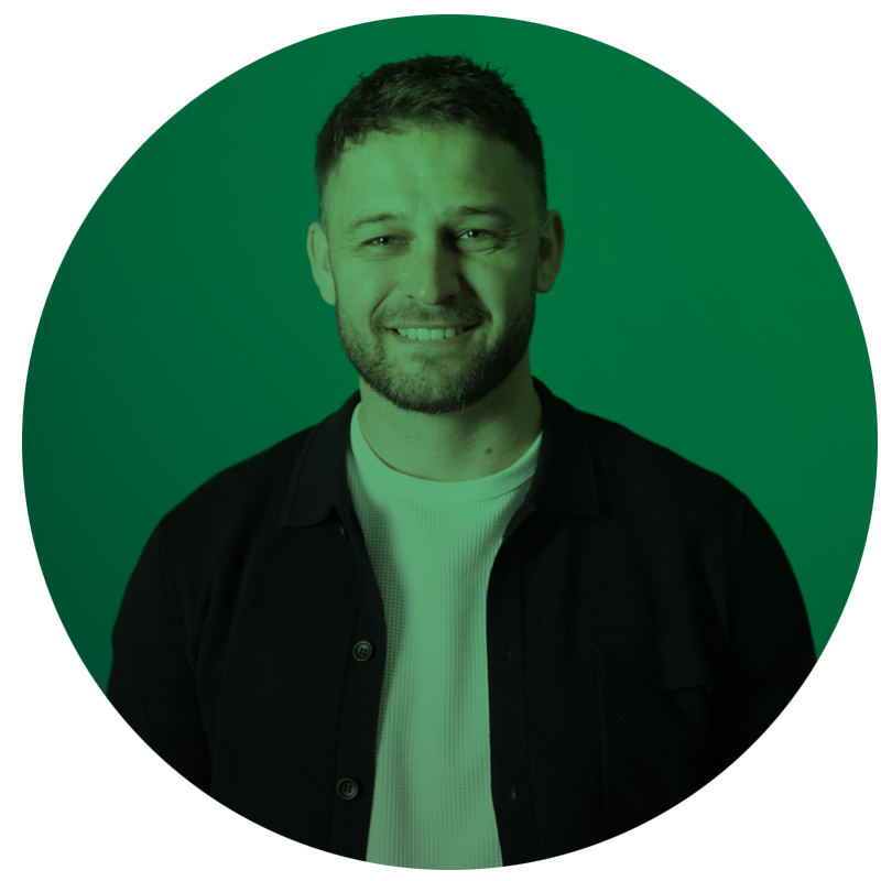 A smiling man with short hair and a beard, wearing a black jacket over a white shirt, standing in front of a green background.