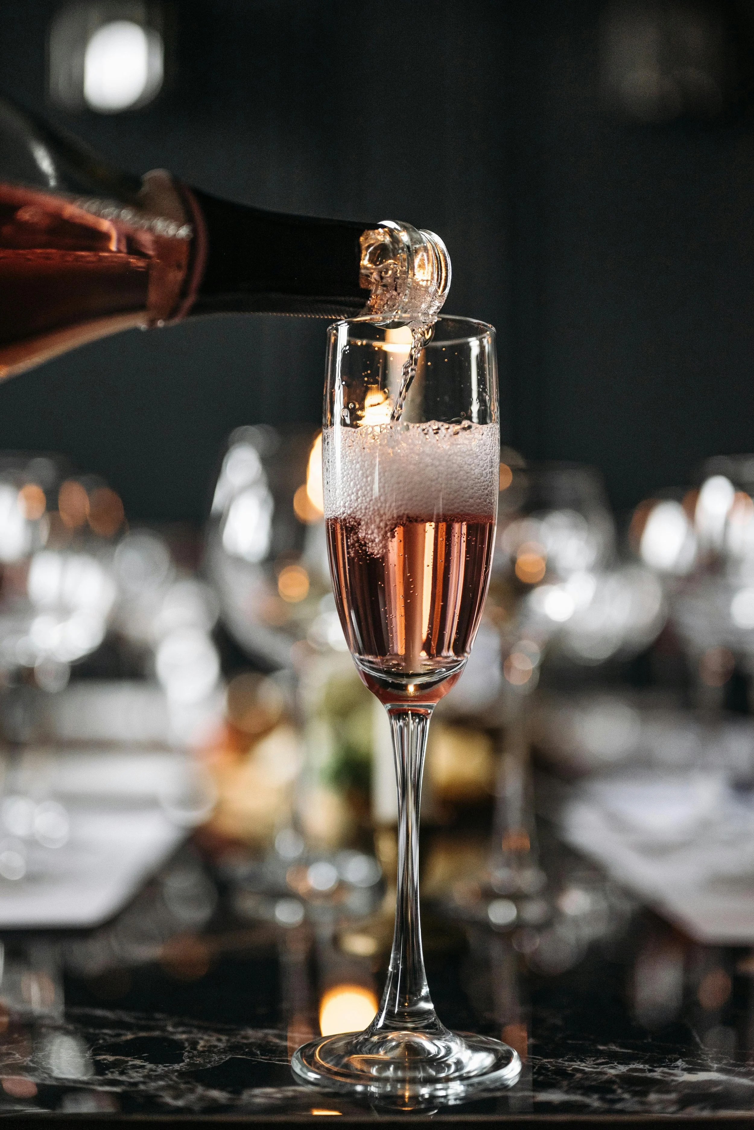 A bottle of pink sparkling wine is being poured into a champagne flute, creating bubbles and foam.