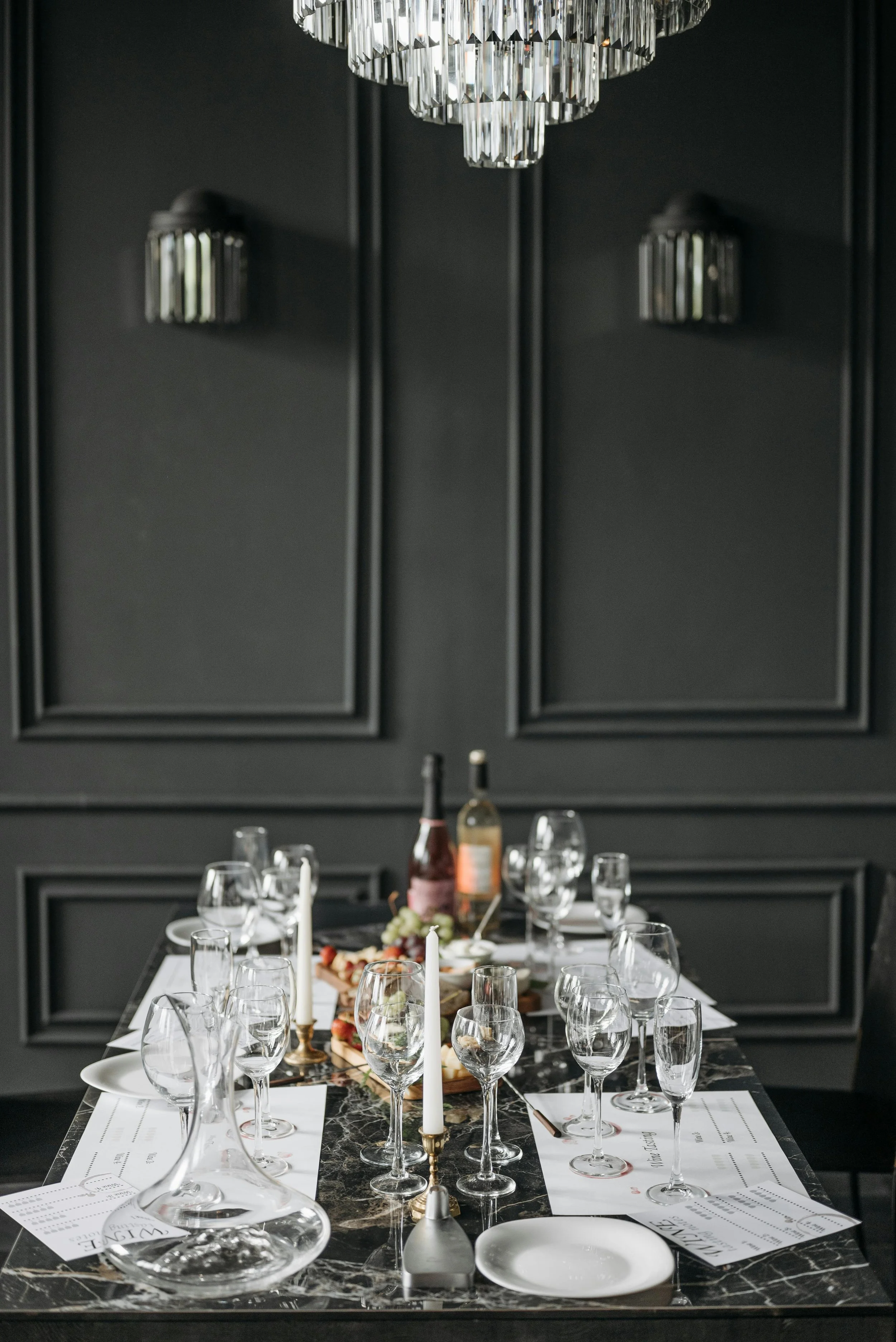 Elegant dining table set with multiple wine glasses, tall white candles, plates, and a charcuterie board, against a dark wall with decorative panels and a crystal chandelier hanging above.