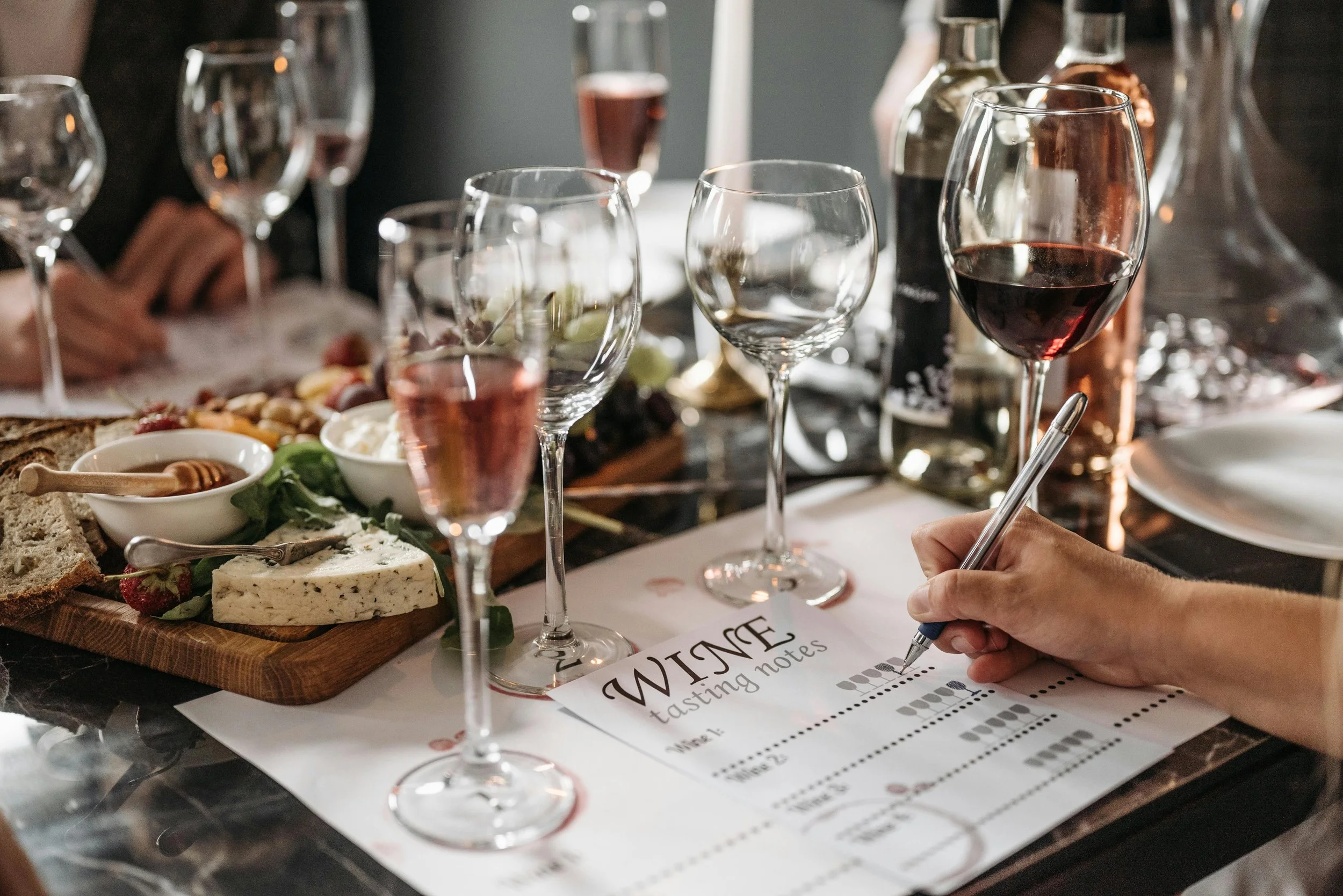 Someone writing tasting notes on a paper titled 'WINE tasting notes' at a crowded wine tasting event, with glasses of red and rosé wine, cheese, bread, and snacks on the table.