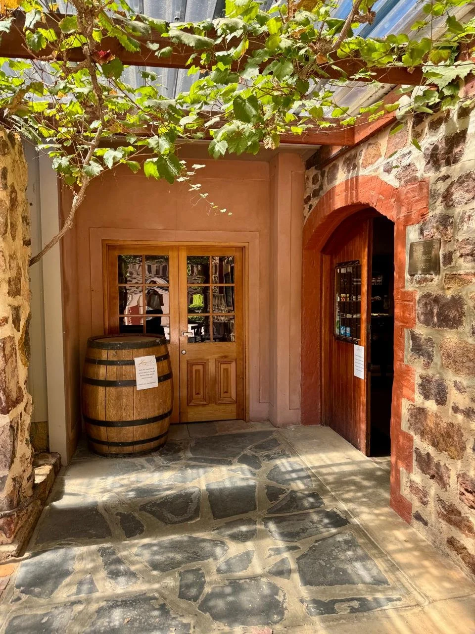 Stone floor leading to rustic wooden door and arched brick doorway, with a wine barrel and leafy vines overhead.