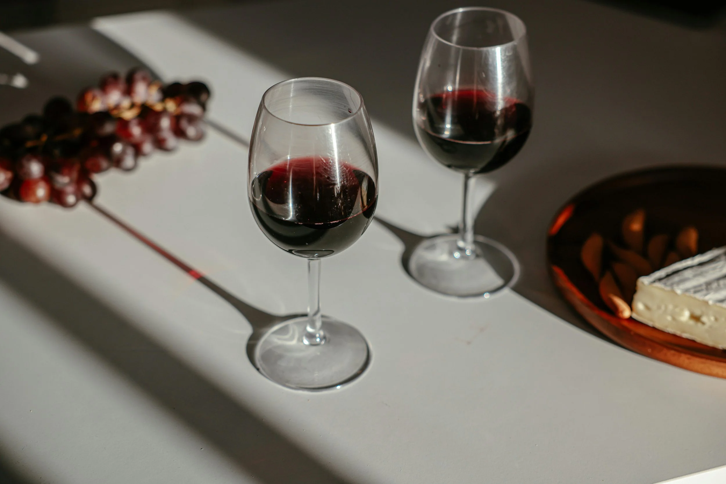 Two glasses of red wine on a white surface next to a plate of cheese and grapes.