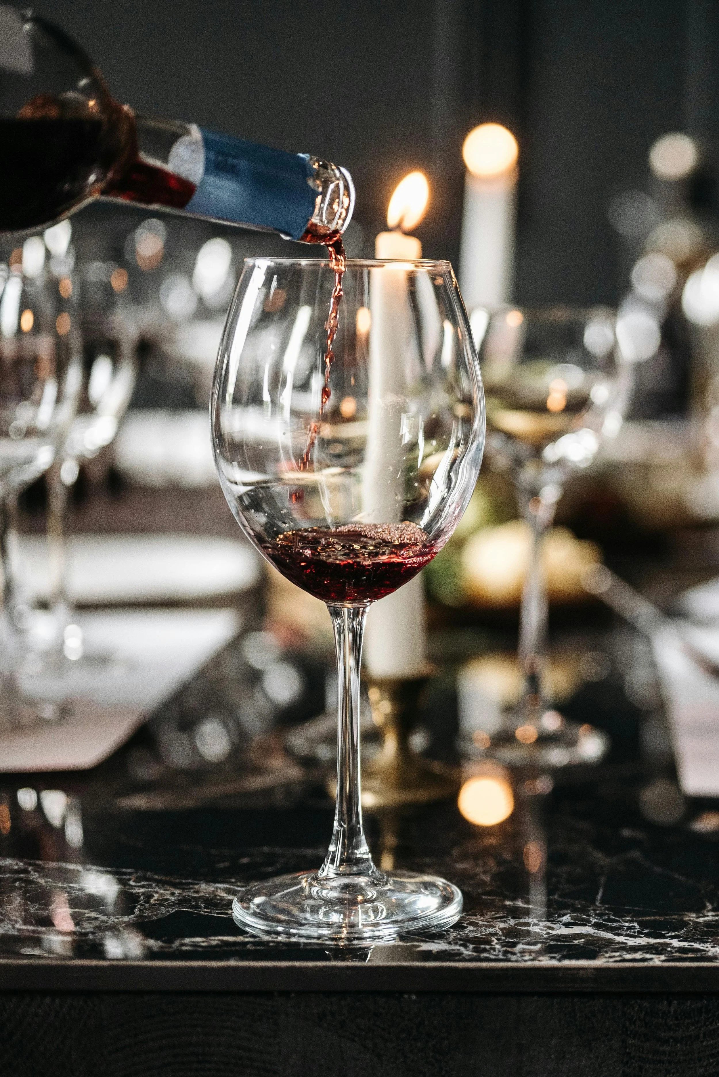 Red wine being poured into a wine glass on a marble table, with candlelight and blurred glassware in the background.