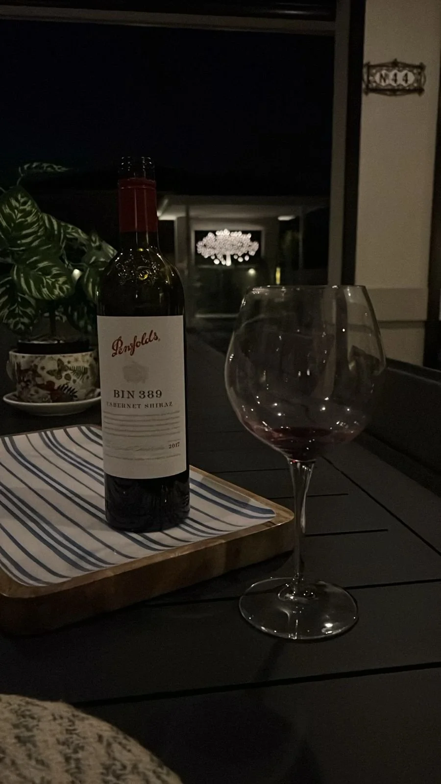 A bottle of Penfolds Bin 389 Cabernet Shiraz wine, partially filled glass of red wine, potted plant, patterned bowl, and striped tray on a black outdoor table at night. Night time wine tasting, drinks by the pool, at home wine tasting.