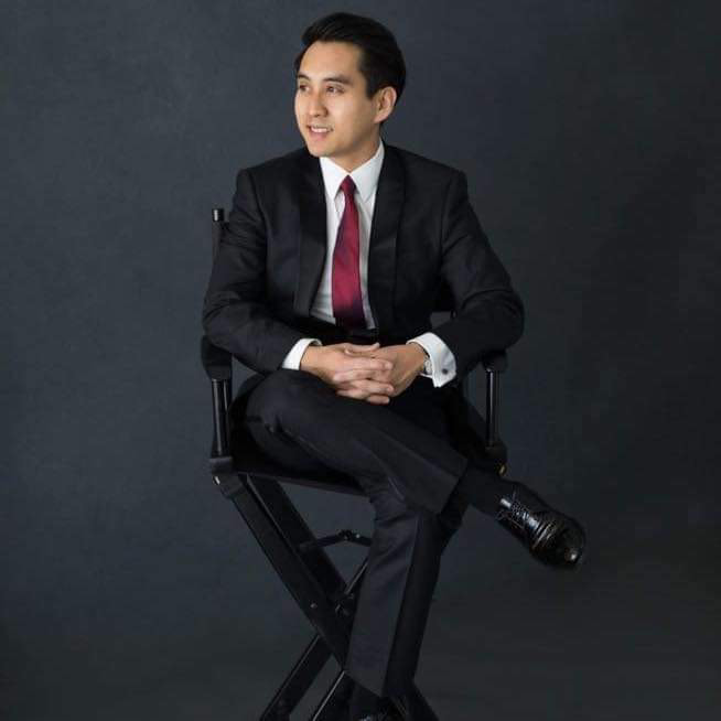 A young man in a black suit and red tie sitting on a director's chair against a dark background, with one leg crossed over the other.
