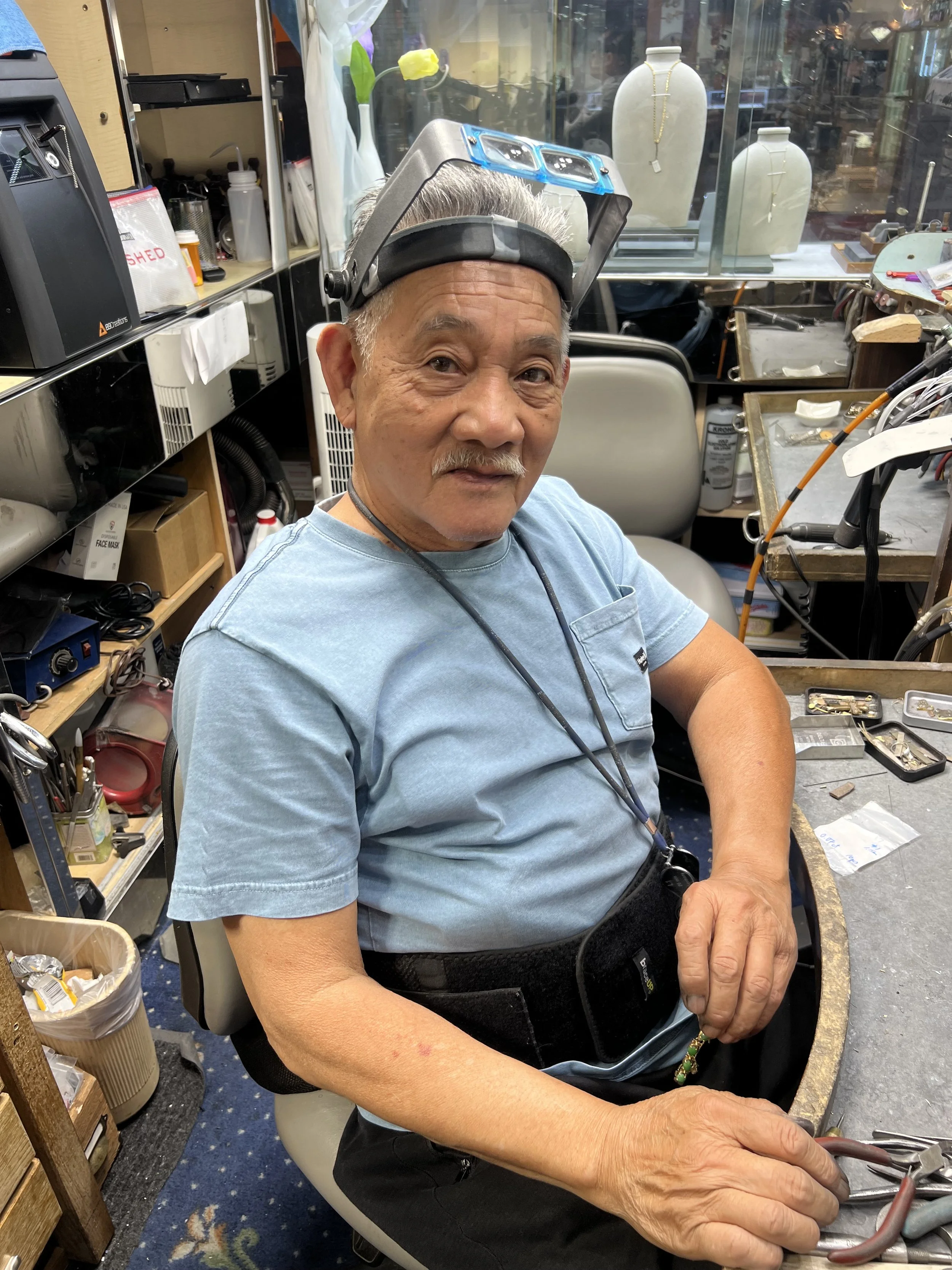 An elderly man wearing a light blue t-shirt, sitting at a workbench in a jewelry workshop, with a magnifying headlamp on his forehead. He is surrounded by tools and jewelry-making supplies, with display cases and mannequin heads with necklaces in the background.