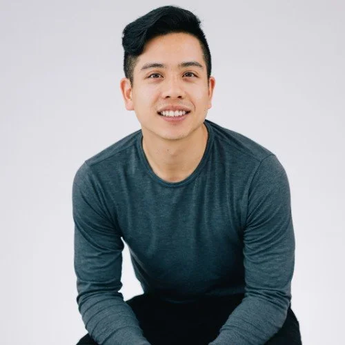 Portrait of a young man with black hair wearing a dark gray long-sleeve shirt, smiling against a plain white background.